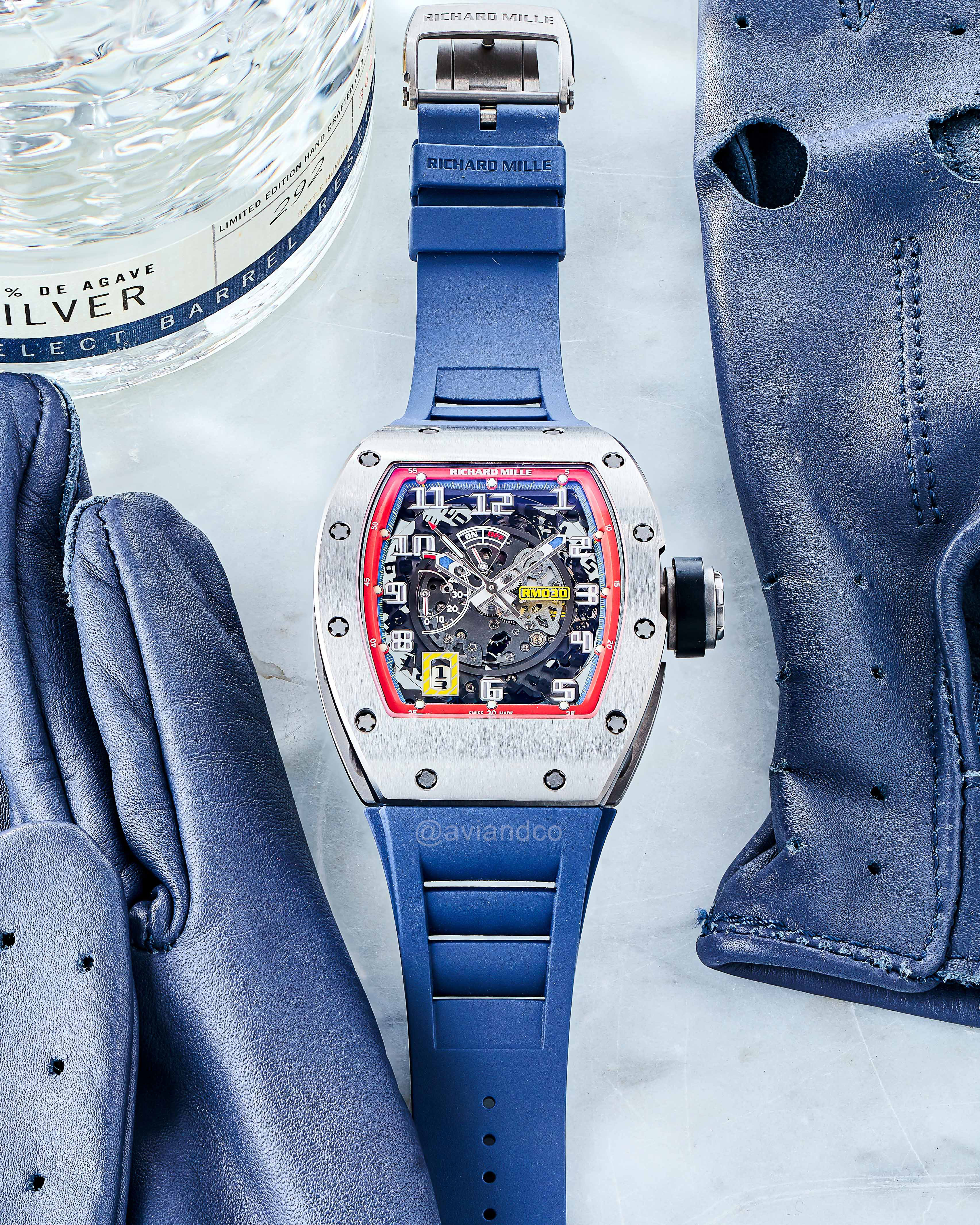 Stainless Steel Bezel with Skeleton Dial and Blue Rubber on a White Marble Tabletop Surrounded by a Bottle of Gin and a Pair of Blue Leather Gloves