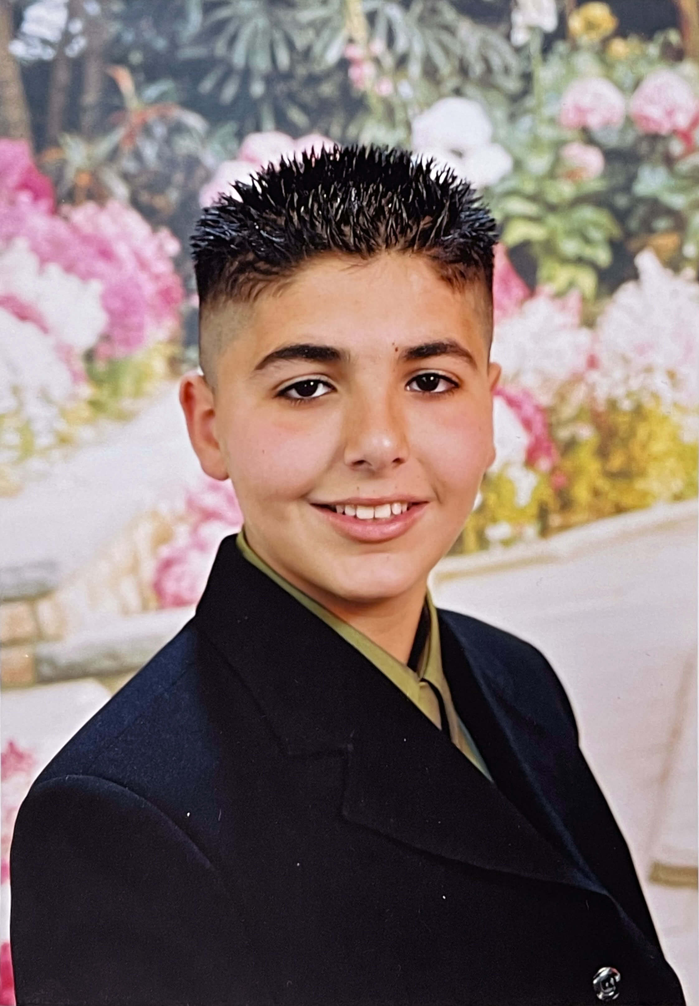 Young Boy From Torso Up Smiling and Wearing a Black Blazer in Front of a Floral Backdrop