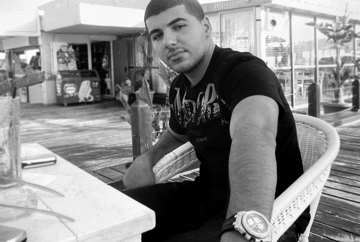 Young Man Sits at a Table Wearing a Black T-shirt with Writing, a Diamond-Set Bezel Around His Wrist, with a Store Front in the Background