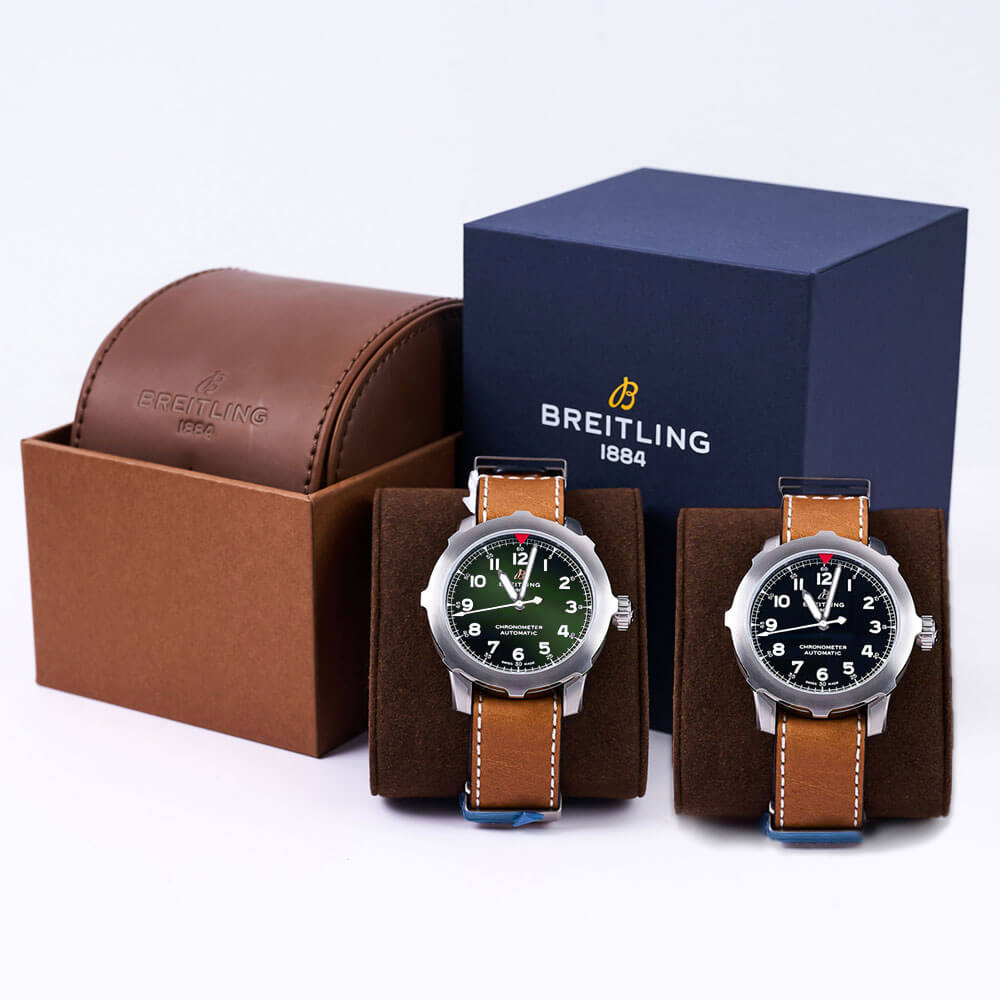 Breitling Aviator Super 8 Light Brown Leather Strap, Sterling Silver Bezel, one with Green Dial and the other with Black Dial, Both Wrapped Around Watch Cushions in front of a Breitling Box
