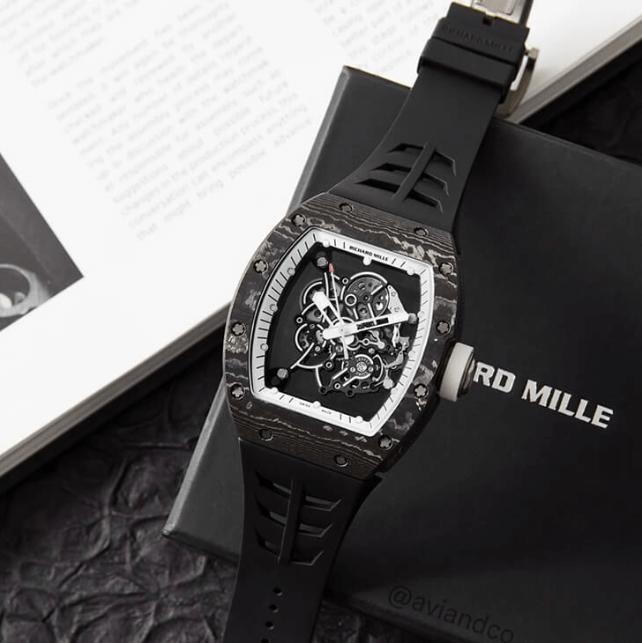 Carbon Bezel with Skeleton Dial and Black Rubber Strap on a Richard Mille Black Box Next to a Book