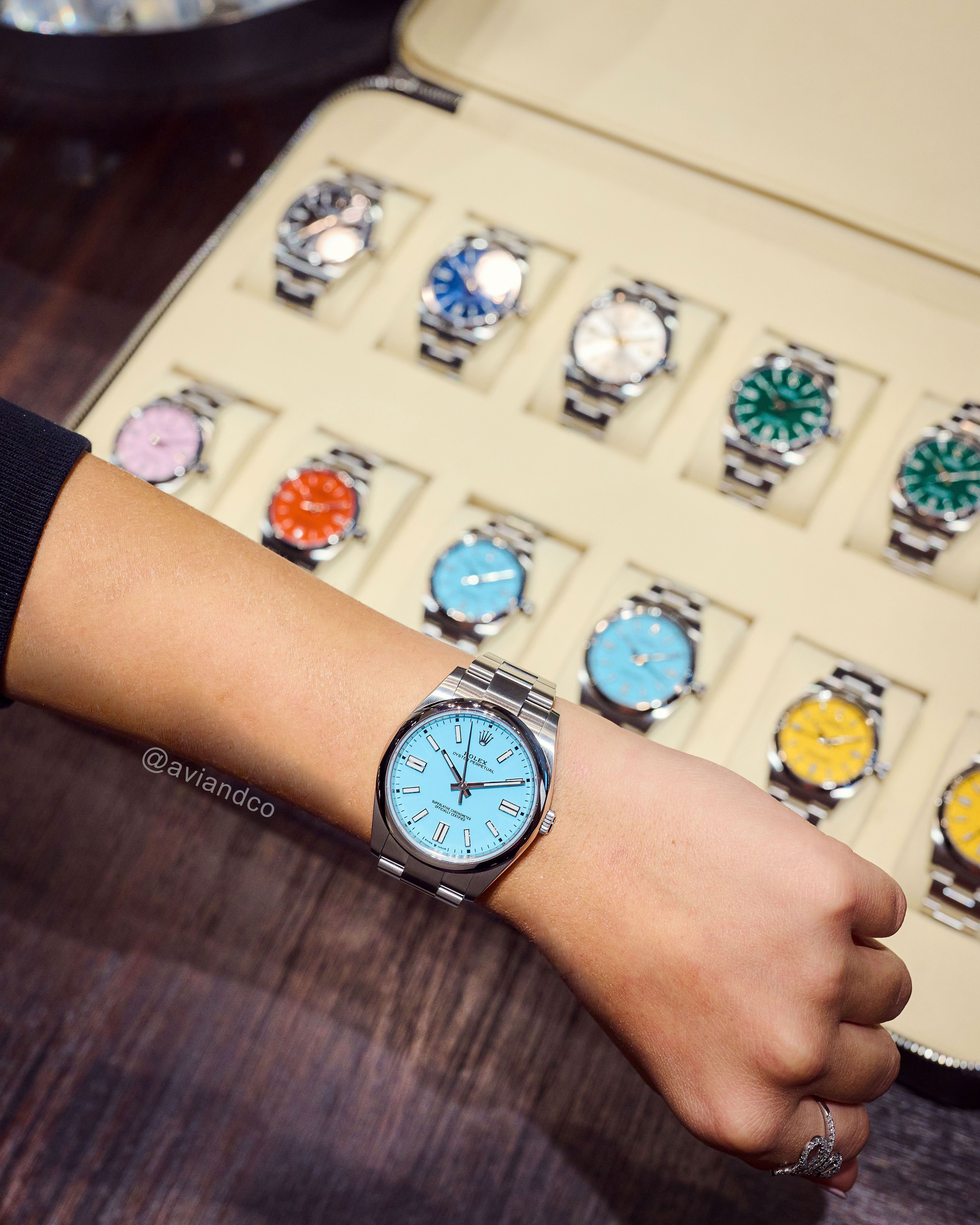 Stainless Steel, Turquoise Index Dial Timepiece on a Woman’s Wrist Above a Watch Case of 12 Colorful Dial Oyster Perpetuals Which are Popular Watch Trends in 2023