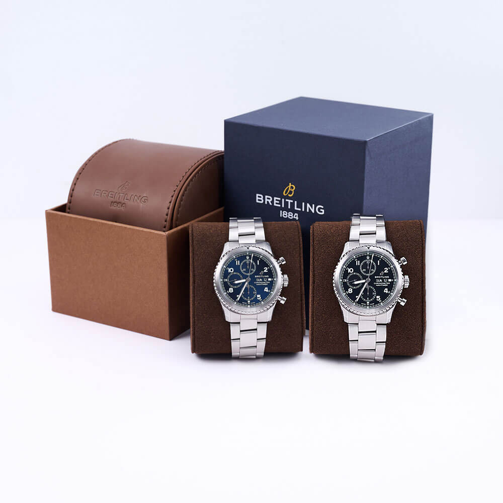 Two Sterling Silver Breitling Navitimer 9 Timepieces, one Blue Dial and the other Black Dial, Both Wrapped Around a Brown Watch Cushion in front of a Breitling Box