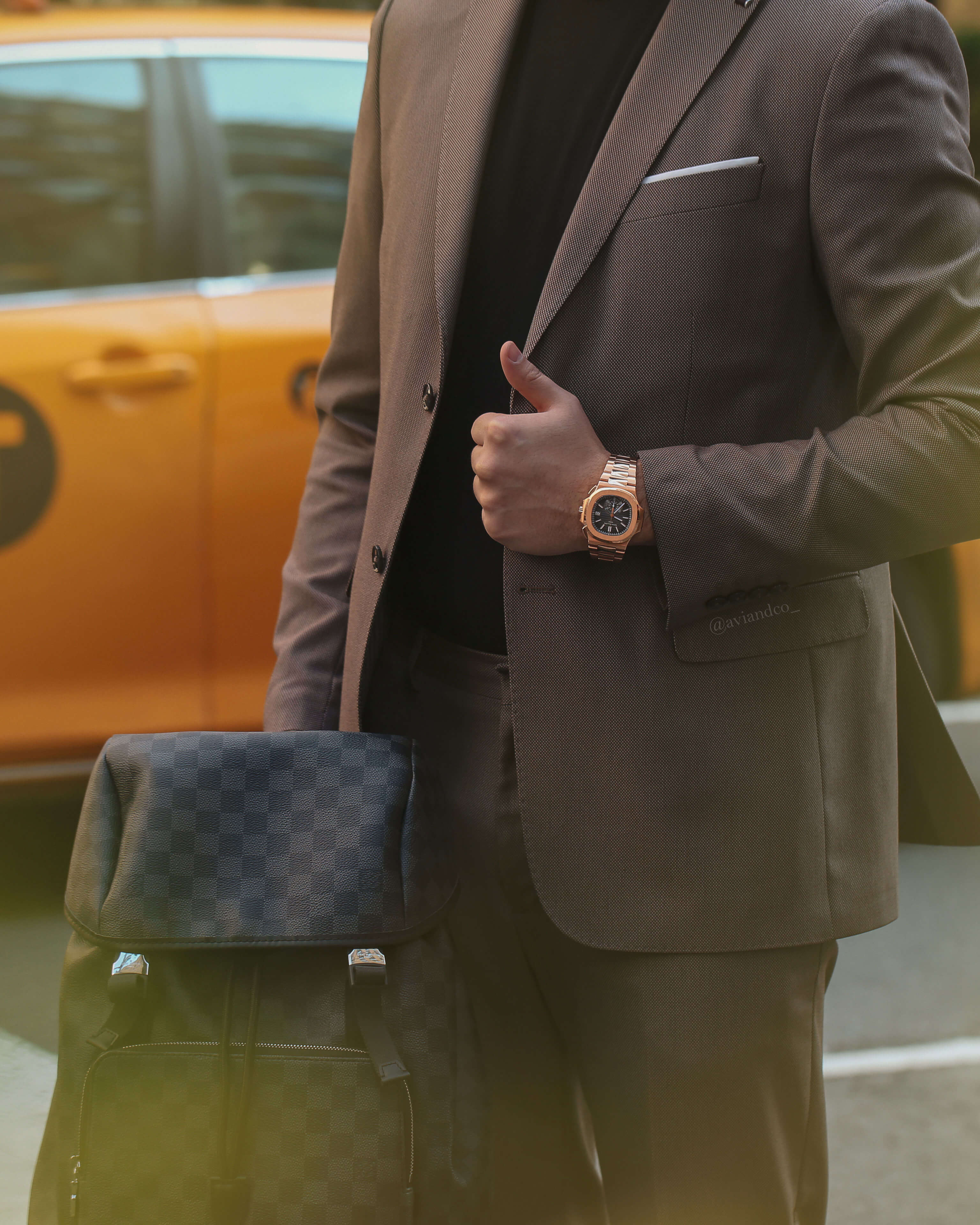 18K Yellow Gold Bezel and Bracelet, Black Dial Timepiece on Man’s Wrist, Wearing a Brown Suit Holding a Checkerboard Backpack in Front of NYC Yellow Taxi