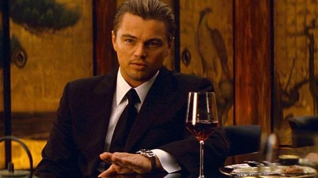 Movie Still from Inception of Leonardo DiCaprio’s Character Wearing a Black Suit, White Button-down Shirt, and Black Tie, Sitting at a Table with a Glass of Red Wine Sporting a Black Leather Strap Timepiece