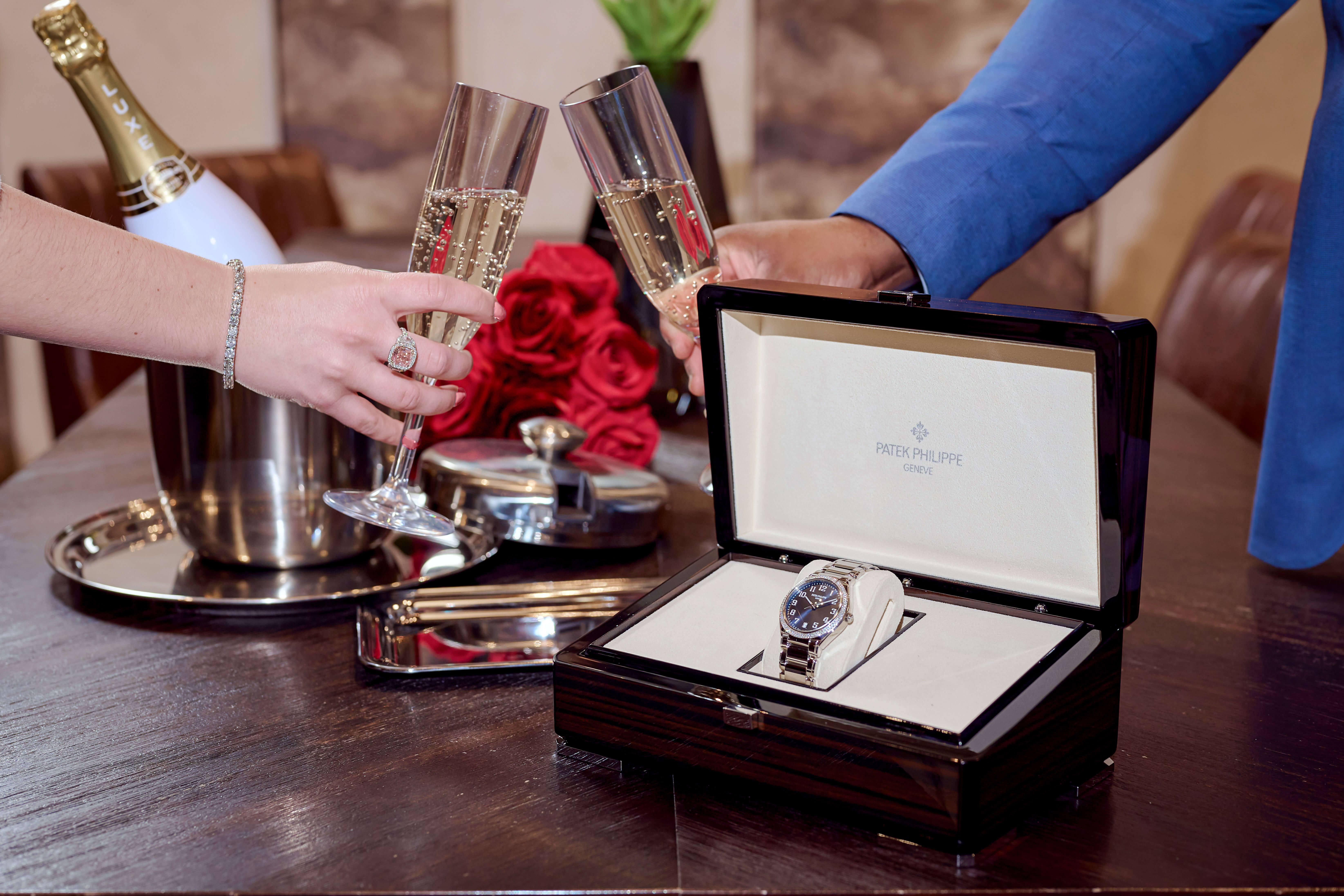 Patek Philippe Twenty~4 Stainless Steel 36 mm Timepiece in a Patek Philippe Dark Brown Watch Box in front of a Woman and Man’s Hand Clinking Champagne Glasses; Champagne Bottle and Roses Adorn the Wooden Table
