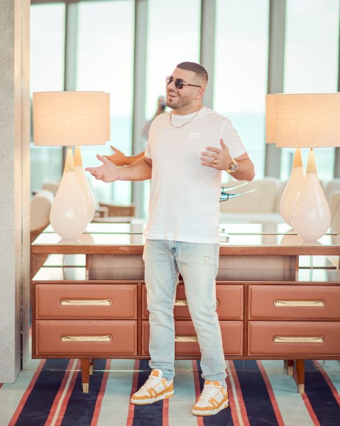 Laughing Young Man Stands In Front of Mirror Wearing White T-shirt, Light Jeans, Orange and White Sneakers, a Watch Around His Wrist, and Sunglasses with Two Cream Lamps and a Wooden Dresser Behind Him