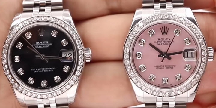 A Left Hand Holds Out Two Rolex Datejusts, One with a Black Dial and the Other With a Pink Dial, Both With Diamond Bezels