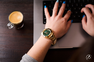 Rolex Daytona, 18K Yellow Gold, Green Dial, 42 mm Case on a Female’s Wrist While She Types on her Laptop that Sits Next to an Espresso.
