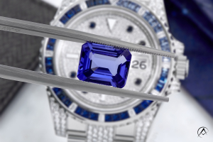 Blue sapphire held with jewelry tongs in front of a watch face with a blue sapphire bezel