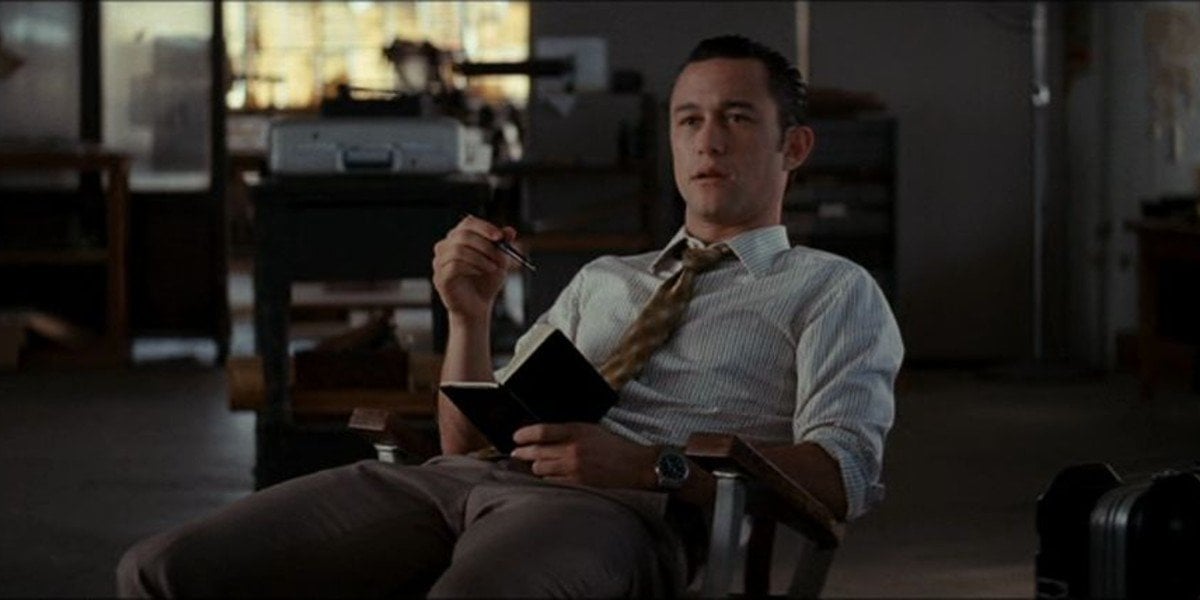 Movie Still from Inception of Joseph Gordon-Levitt’s Character Sitting on a Chair in an Office Setting, Holding a Black Notebook and Sporting a White Button Down Shirt, Brown Tie, and IWC Luxury Timepiece