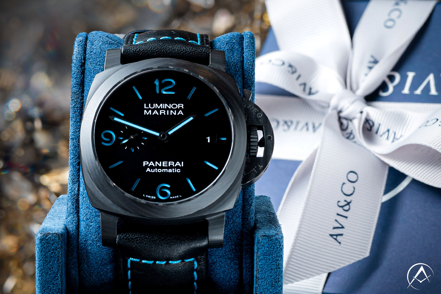 Panerai Luminor Marina Watch with 44 mm Carbon Case, Black Dial with Blue Luminous Hour Markers, and Black Leather Bracelet Displayed on a Blue Velvet Watch Cushion Beside a Blue and White Avi & Co. Gift Box.