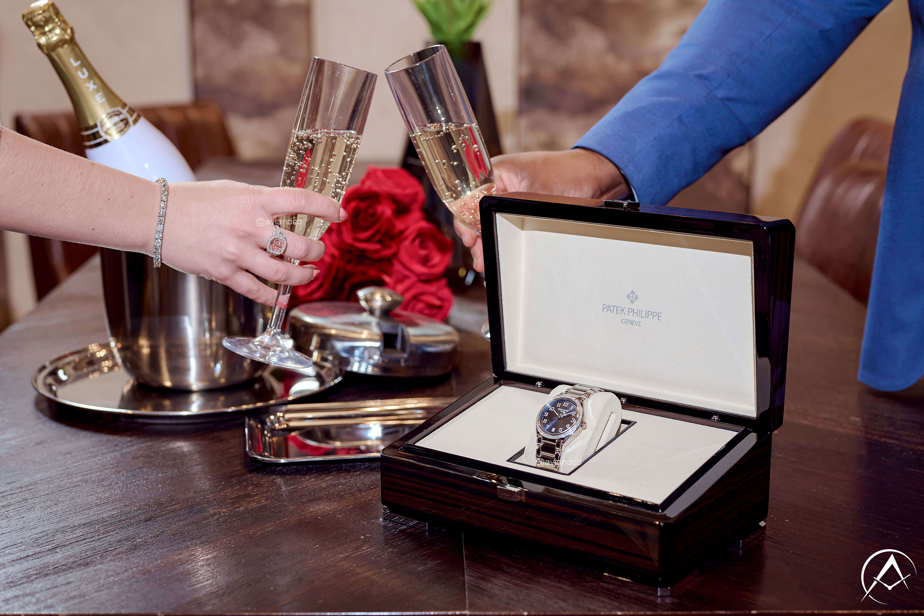 Patek Philippe Twenty~4 Stainless Steel 36 mm Timepiece in a Patek Philippe Dark Brown Watch Box in Front of a Woman and Man’s Hand Clinking Champagne Glasses. Champagne Bottle and Roses Adorn the Wooden Table.