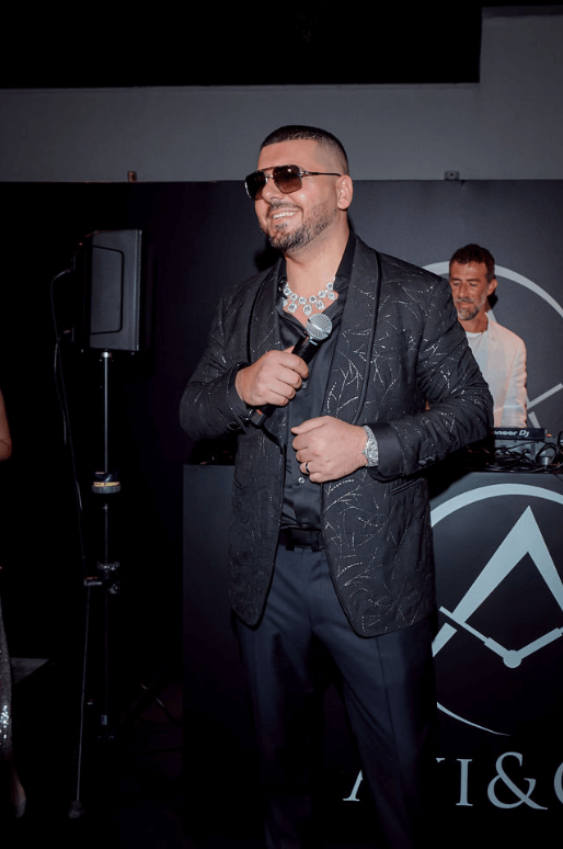 CEO of Avi & Co. Wears Black Suit, The Crown Chain Necklace, and the Avi & Co. Limited Edition Watch Around His Wrist While Holding a Microphone in Front of the DJ.