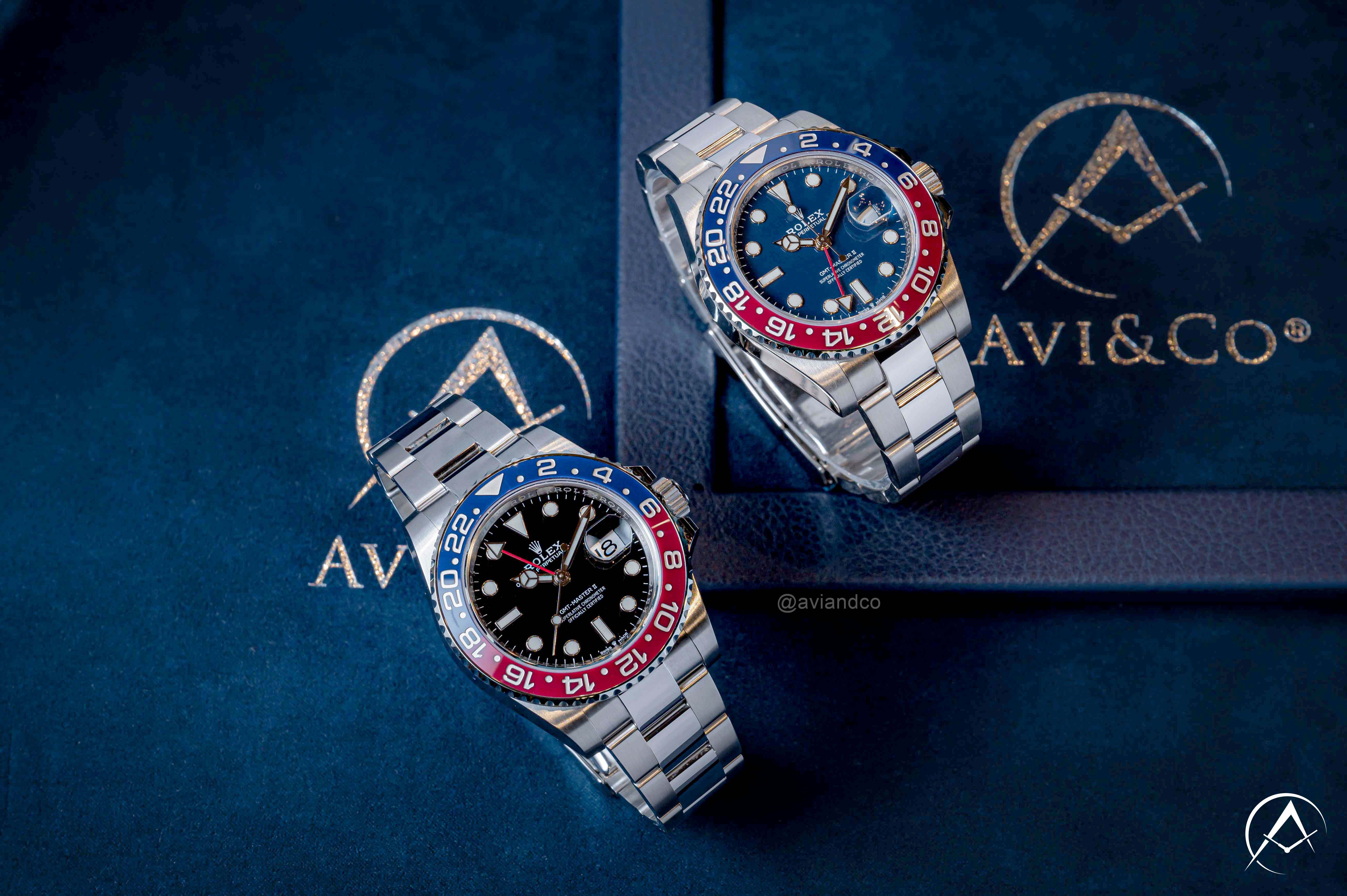 Two Similar Luxury Timepieces with Red and Blue Pepsi Bezels, 40 mm Case Sizes, and Different Materials Displayed on Clear C-Rings Above a Navy Blue Avi & Co. Watch Pad.