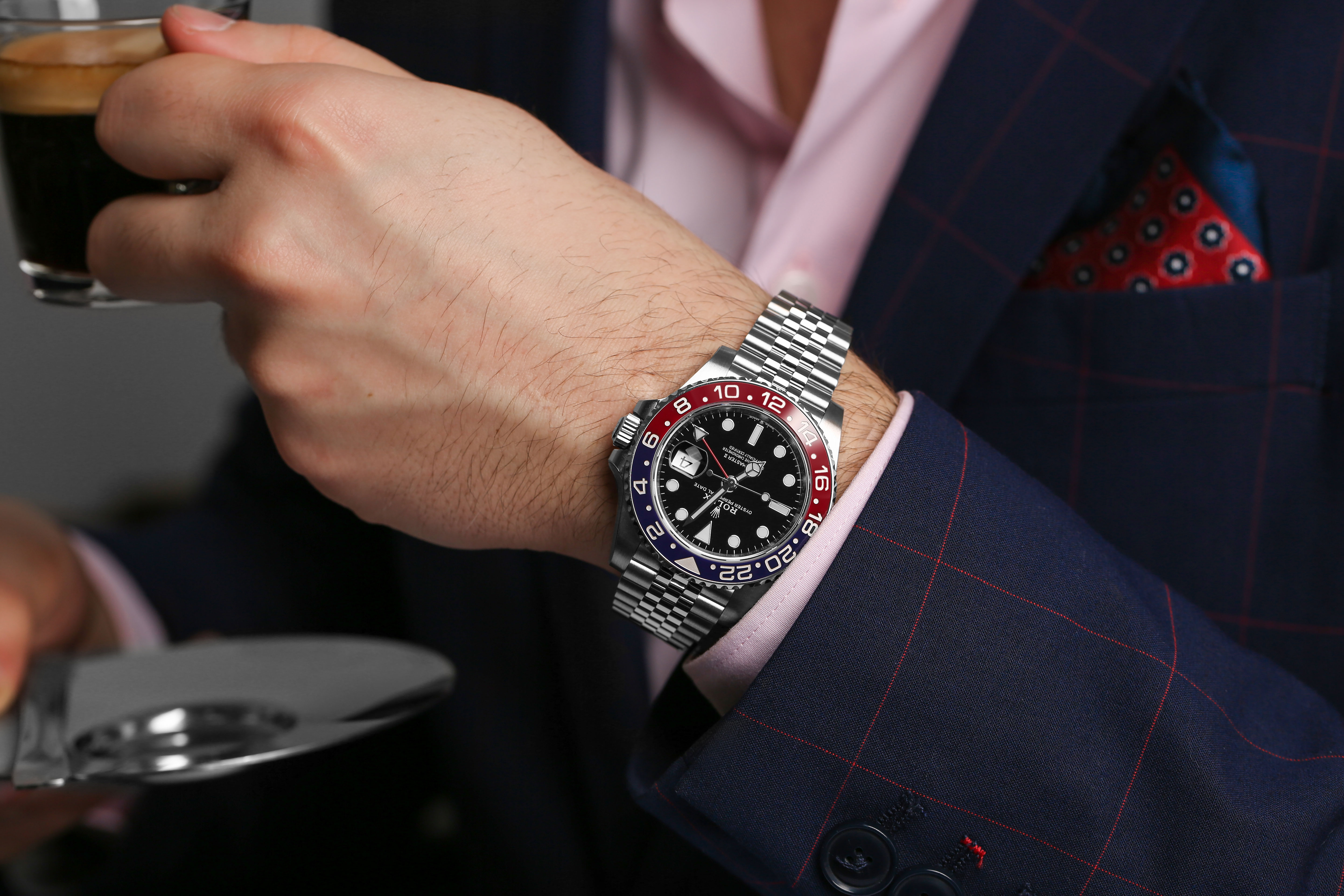 Stainless Steel Timepiece with Black Dial and Two Tone Bezel Sits on a Man’s Wrist who is Drinking Coffee and Wearing a Navy Blue Shirt with Pink Button Down Shirt.