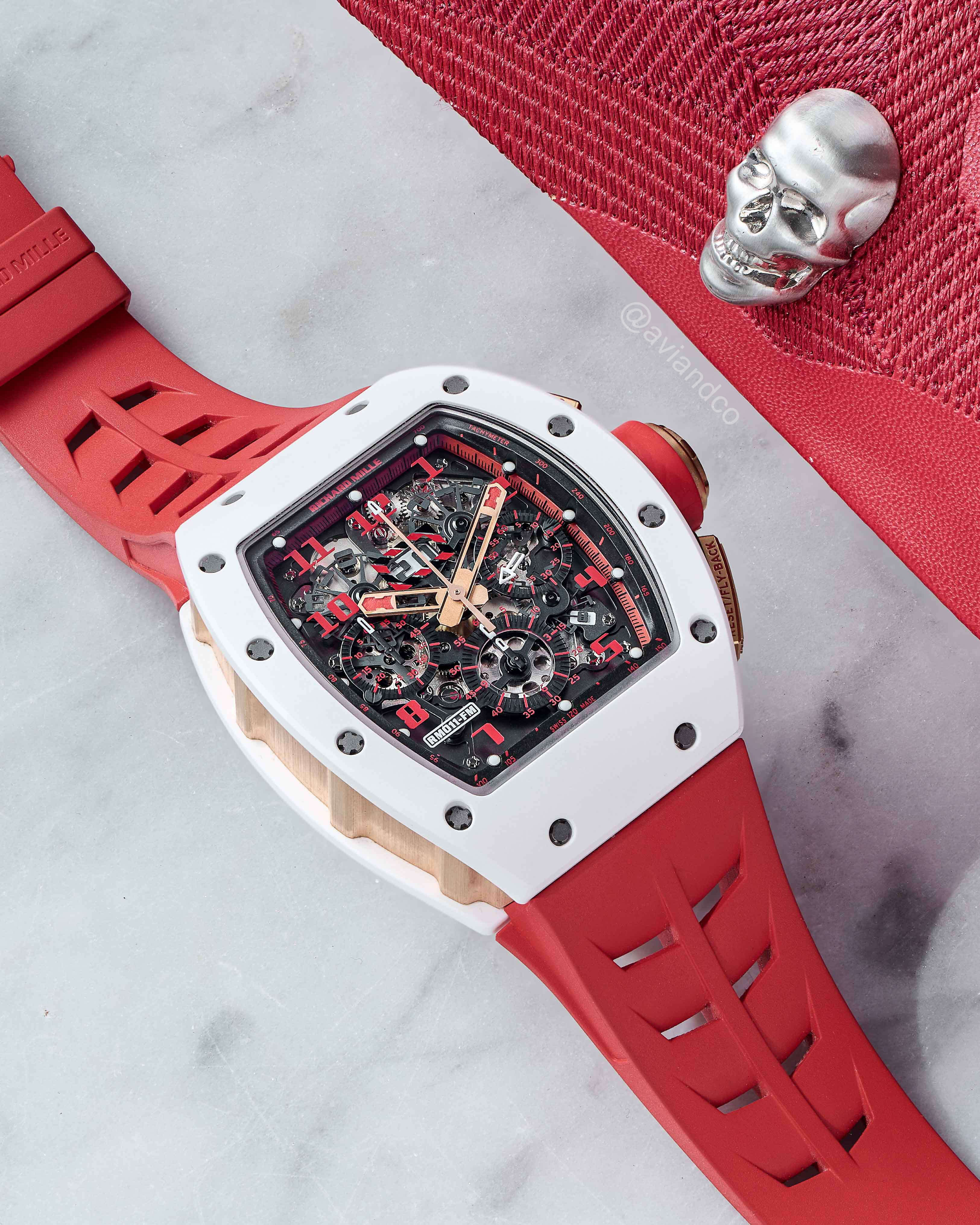 Red Rubber Strap Richard Mille Timepiece with White Bezel and Red Hour Markers Laid Out on White Marble Tabletop.