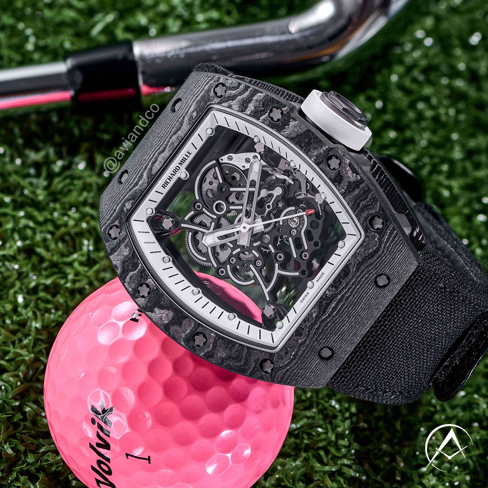 Black Carbon TPT Timepiece with Skeleton Dial Lying Beside a Pink Golf Ball and a Silver Golf Club on Grass.
