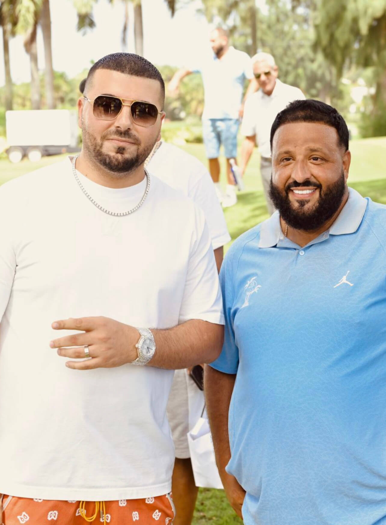 Avi of Avi & Co. Wears a White T-Shirt, Diamond Chain, Sunglasses, and Diamond Timepiece, Standing Beside DJ Khaled in a Blue Collared T-Shirt on a Golf Course.