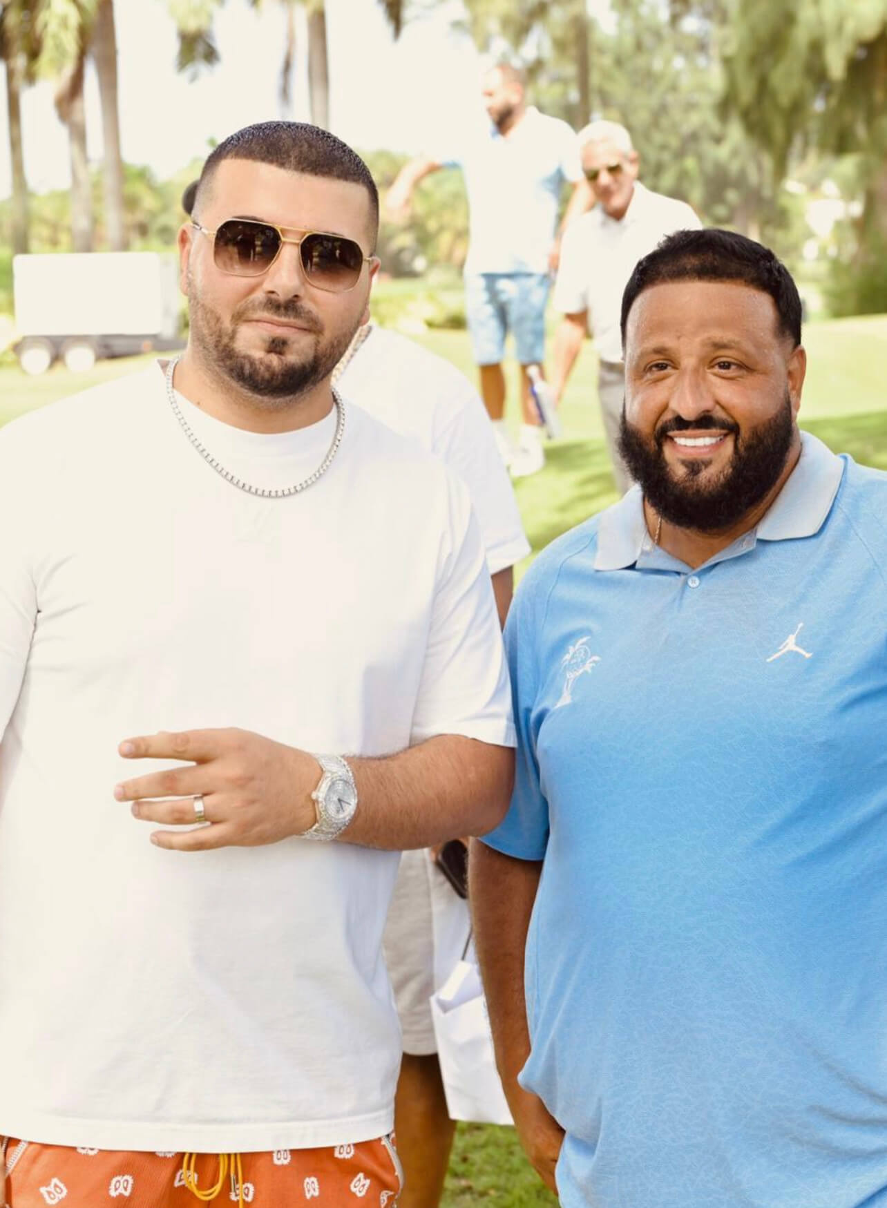 Avi Hiaeve of Avi & Co. Wears a White T-Shirt, Diamond Chain, Sunglasses, and Diamond Timepiece, Standing Beside DJ Khaled in a Blue Collared T-Shirt on a Golf Course.