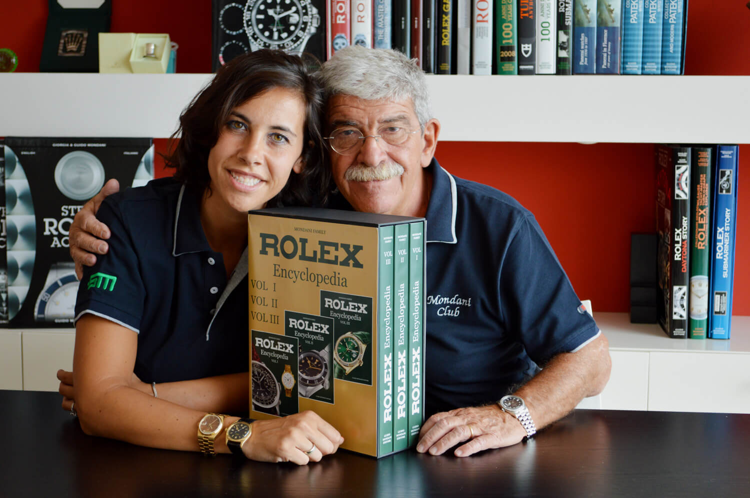 Giorgia and Guido Mondani Sport Watches as they Display The Rolex Encyclopedia in front of a Bookshelf.