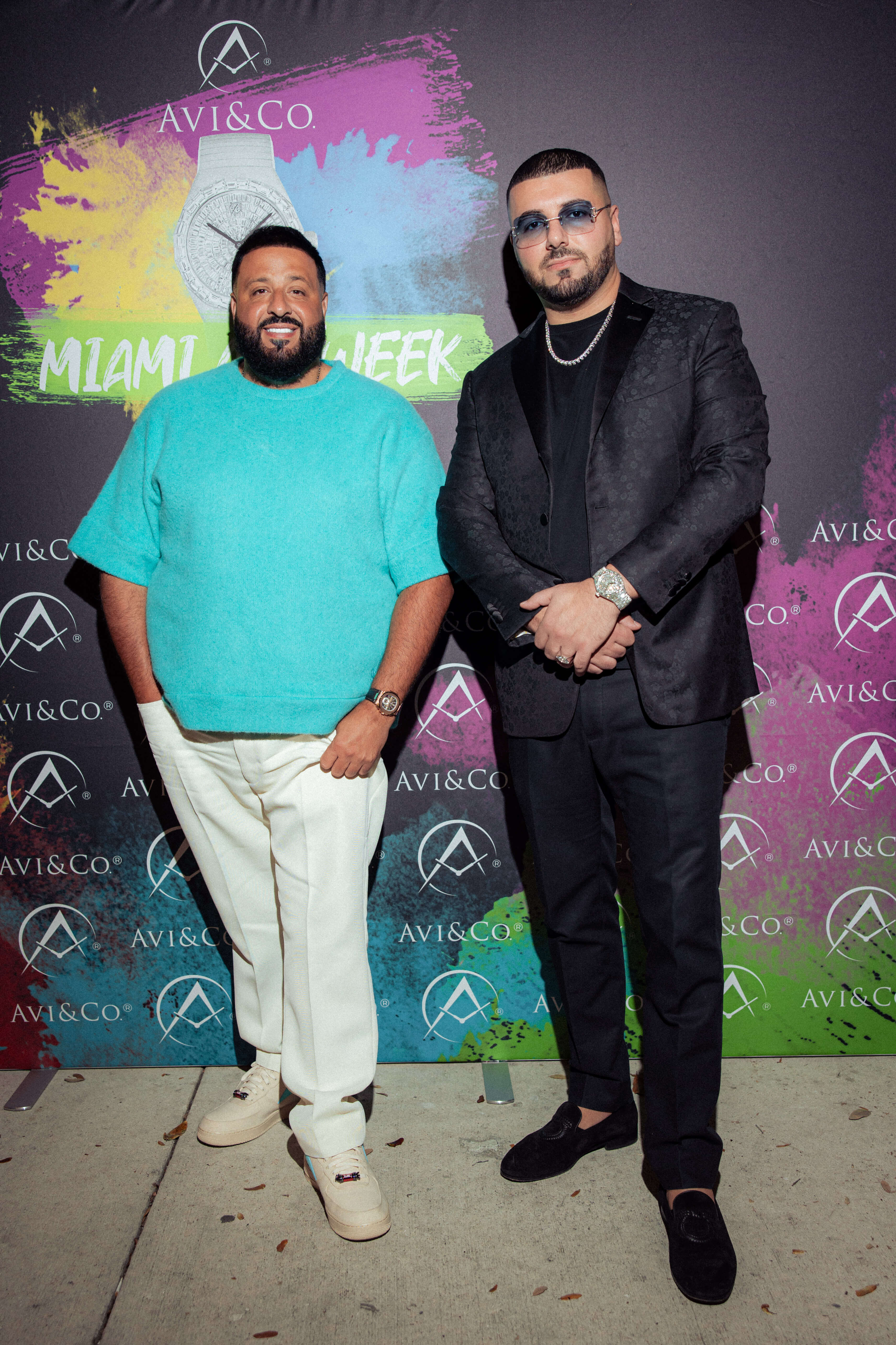 Two Men Stand in Front of Avi & Co.'s Step and Repeat. Left: Man Wears Blue T-Shirt and White Pants. Right: Man Wears Black Suit with Diamond-Pave Chain. Both Wear Luxury Timepieces.