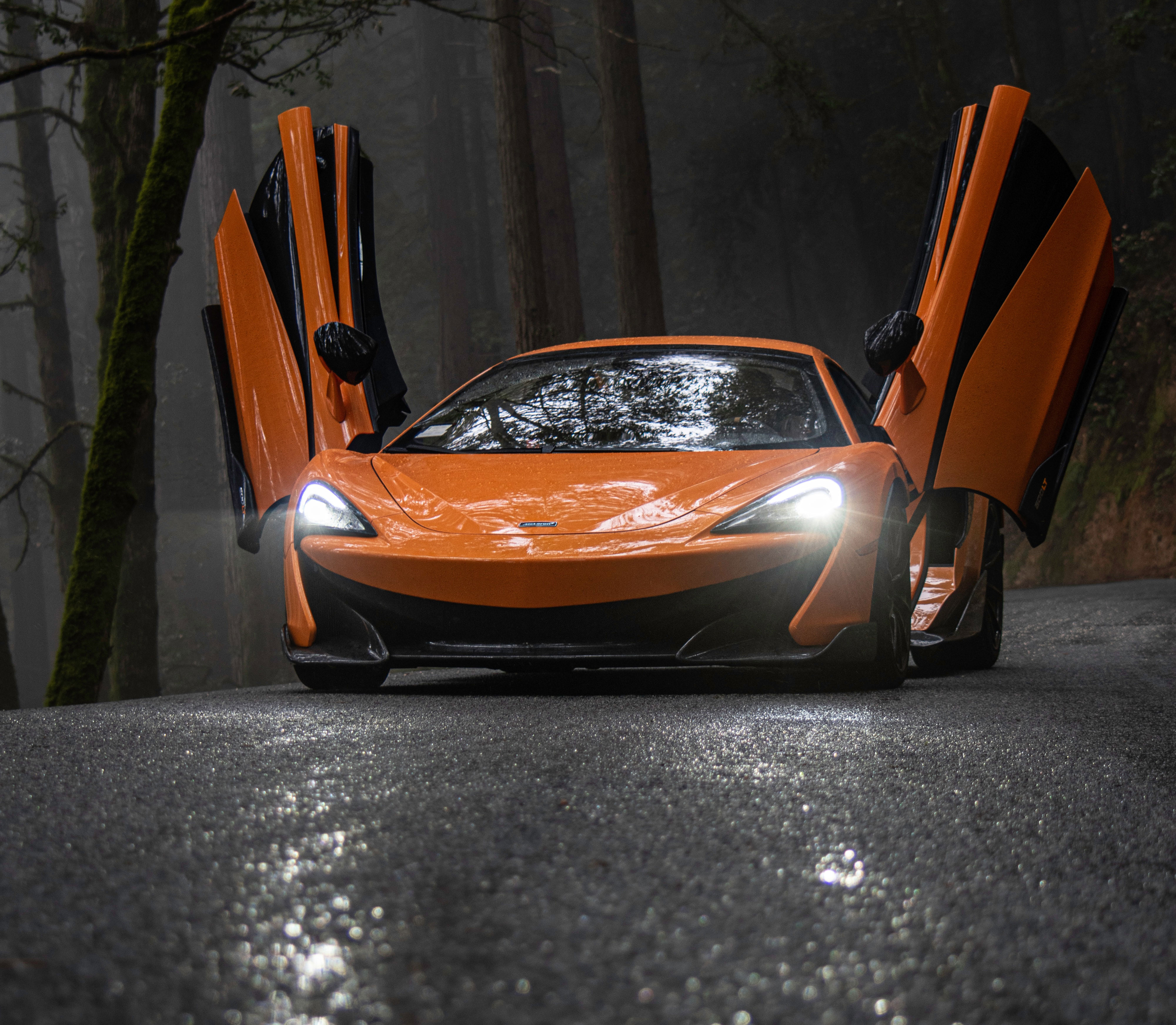 Orange Sports Car with Lights on and Car Doors Open to the Sky in a Dark Wooded Location.