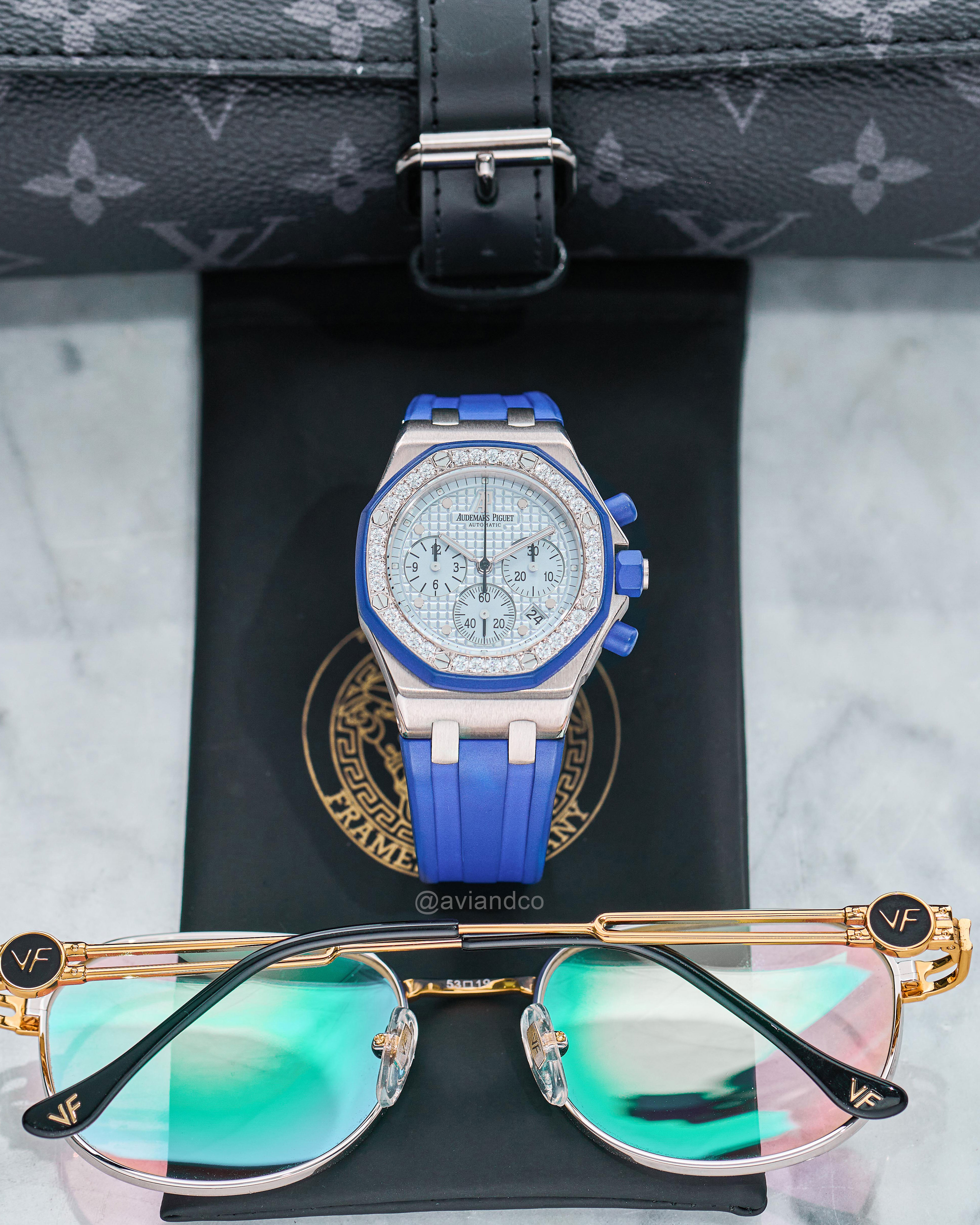 Diamond-Pave Bezel Timepiece with Blue Rubber Strap Beside a Louis Vuitton Bag and Iridescent Sunglasses.