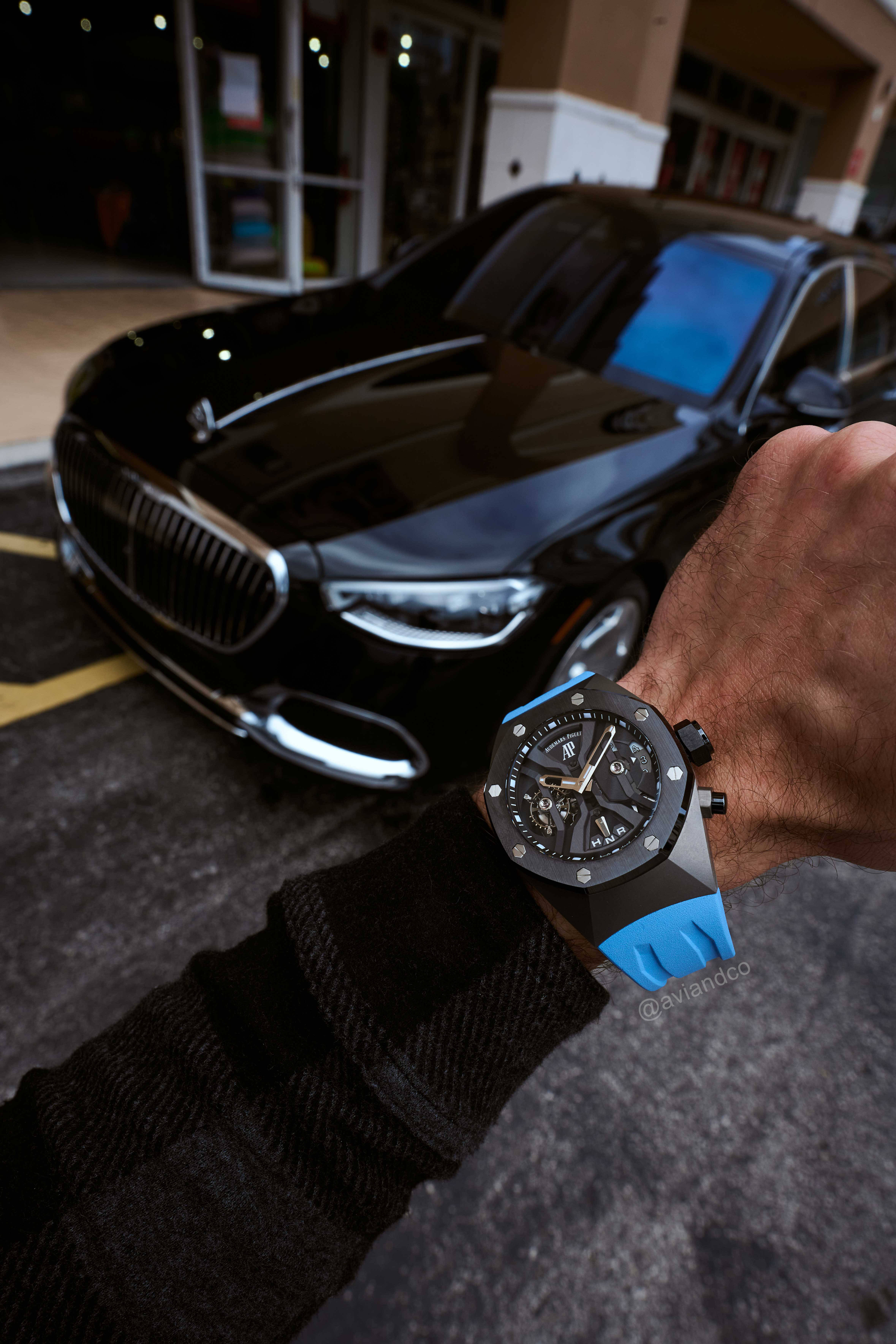 Openworked Dial with Black Bezel Timepiece on a Blue Rubber Strap on a Man’s Wrist in Front of a Black Luxury Car.