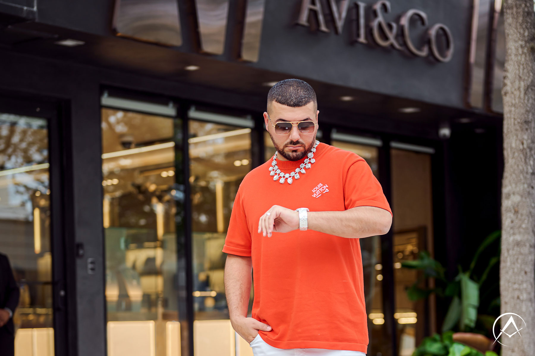 Man Wears Louis Vuitton Orange Shirt, White Pants, and Orange Sneakers with Diamond Watch and Necklace in Front of Avi & Co.’s Miami Boutique.