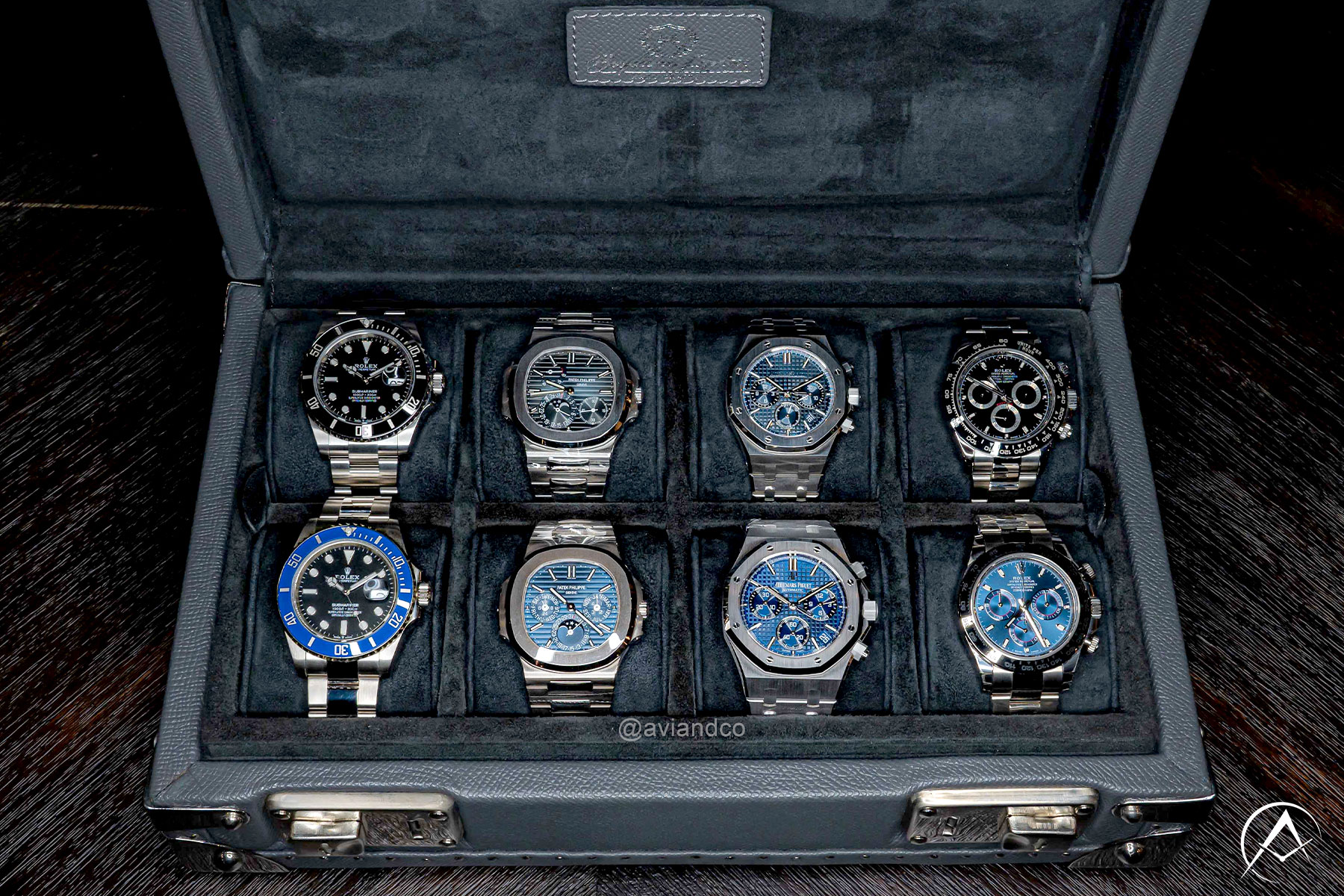 Navy Blue Avi & Co. Watch Case Featuring Four Stainless Steel Luxury Timepieces on the Top Row and Four White Gold Luxury Timepieces on the Bottom Row. They are a Mixture of Rolex, Audemars Piguet, and Patek Philippe watches.