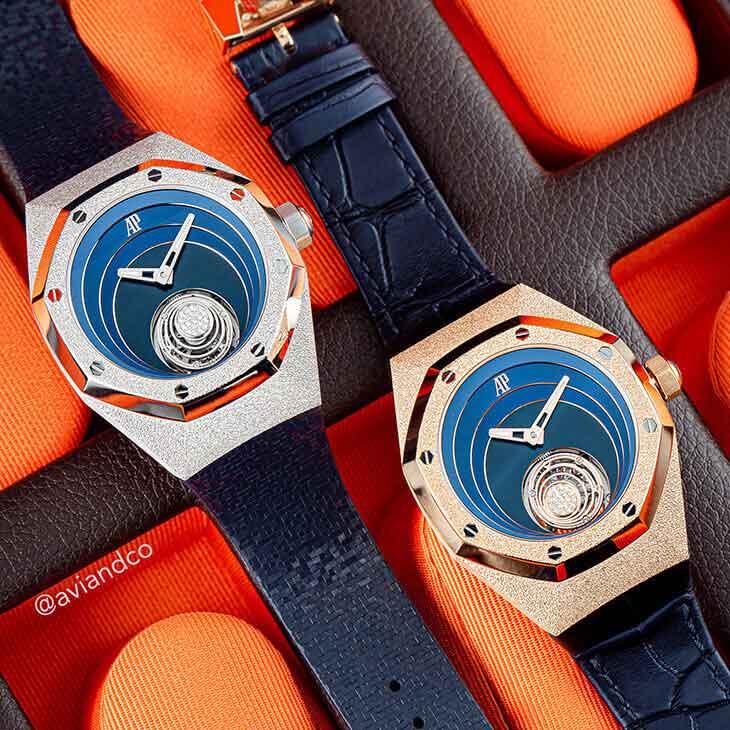Two Blue Dial Timepieces with a White and Gold Bezel Lay Open on an Orange Background with Navy Leather Straps.