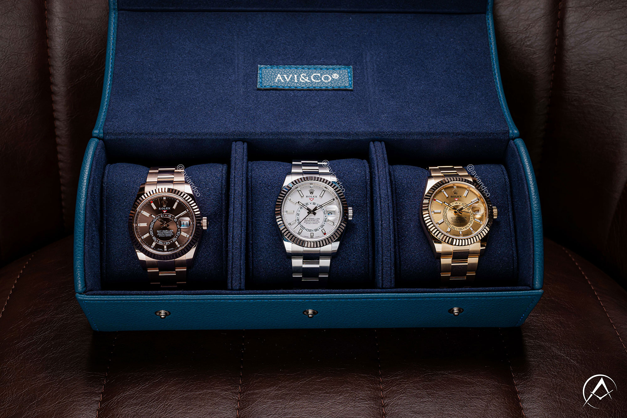 Three Luxury Timepieces Displayed in Navy Avi & Co. Watch Roll. From Left to Right: 18K Rose Gold Chocolate Index Dial, Stainless Steel White Dial, and 18K Yellow Gold Champagne Dial, All with Dual Time Zones.
