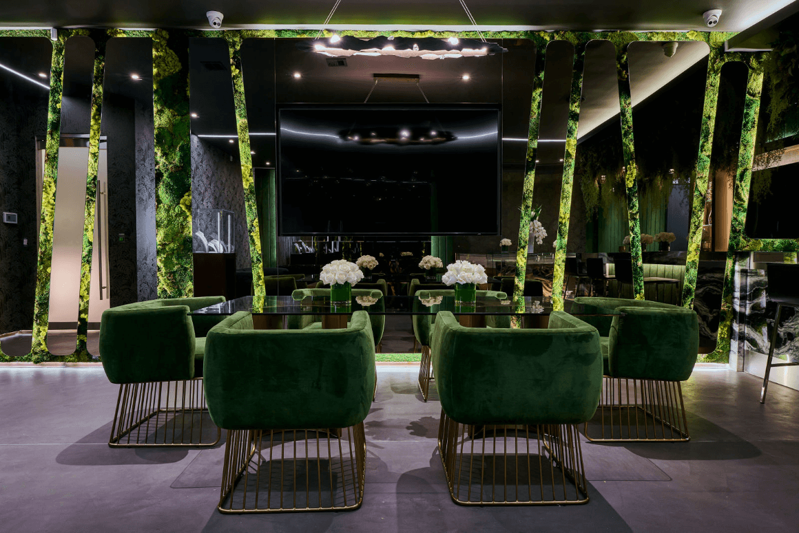 Showroom Decorated with Glass Table with Six Green Velvet Chairs, a Flatscreen TV, and a Bar.