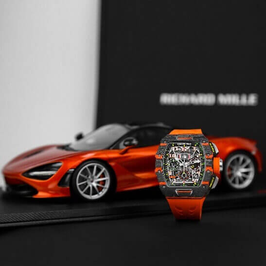 Carbon 44 mm Timepiece Assembled with Quartz TPT Bezel and Orange Rubber Strap Displayed on a C-Ring in Front of an Orange McLaren Car.