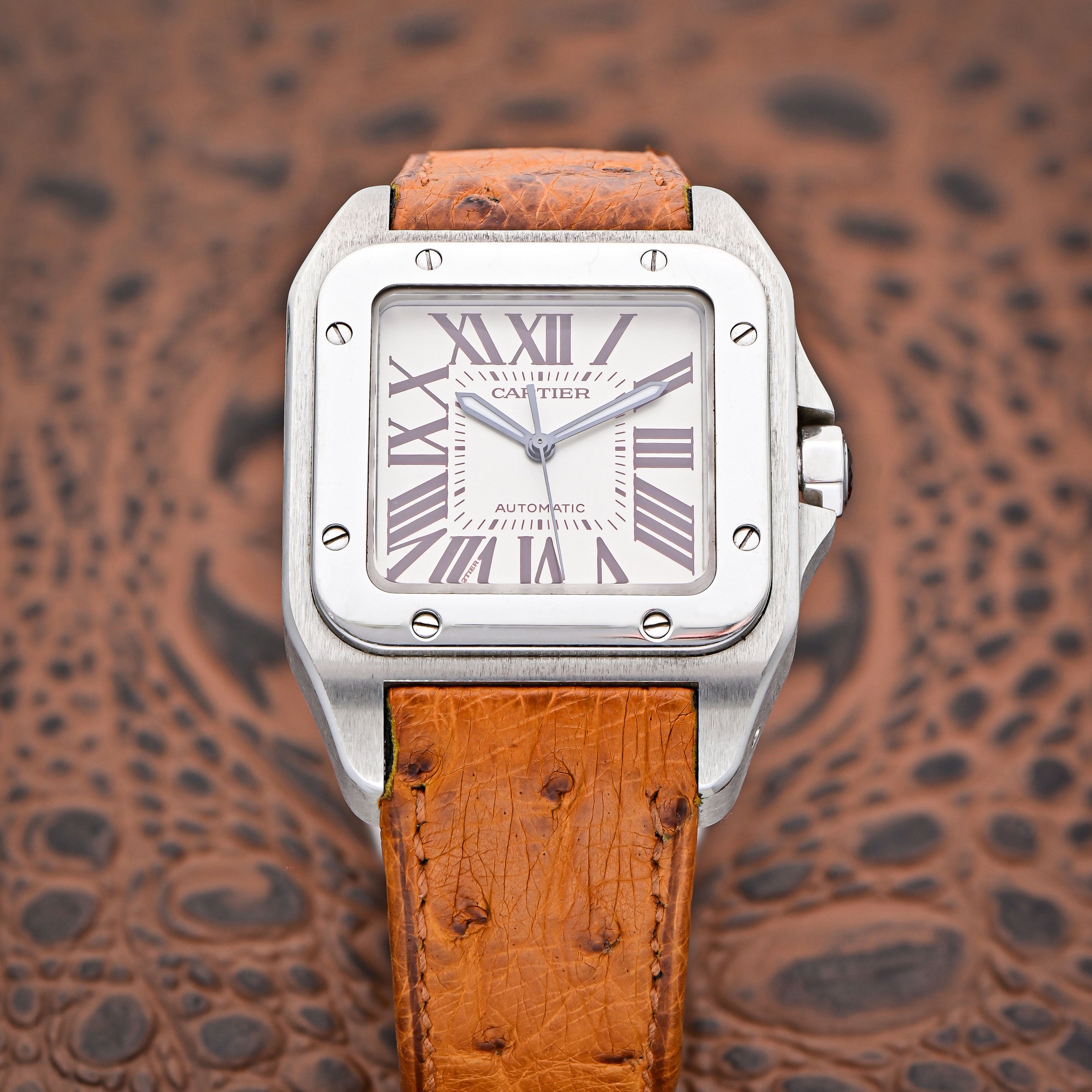 White Dial with Black Roman Numerals on a Square-Shaped Case with a Light Brown Leather Strap on a Brown and Black Leather Background.