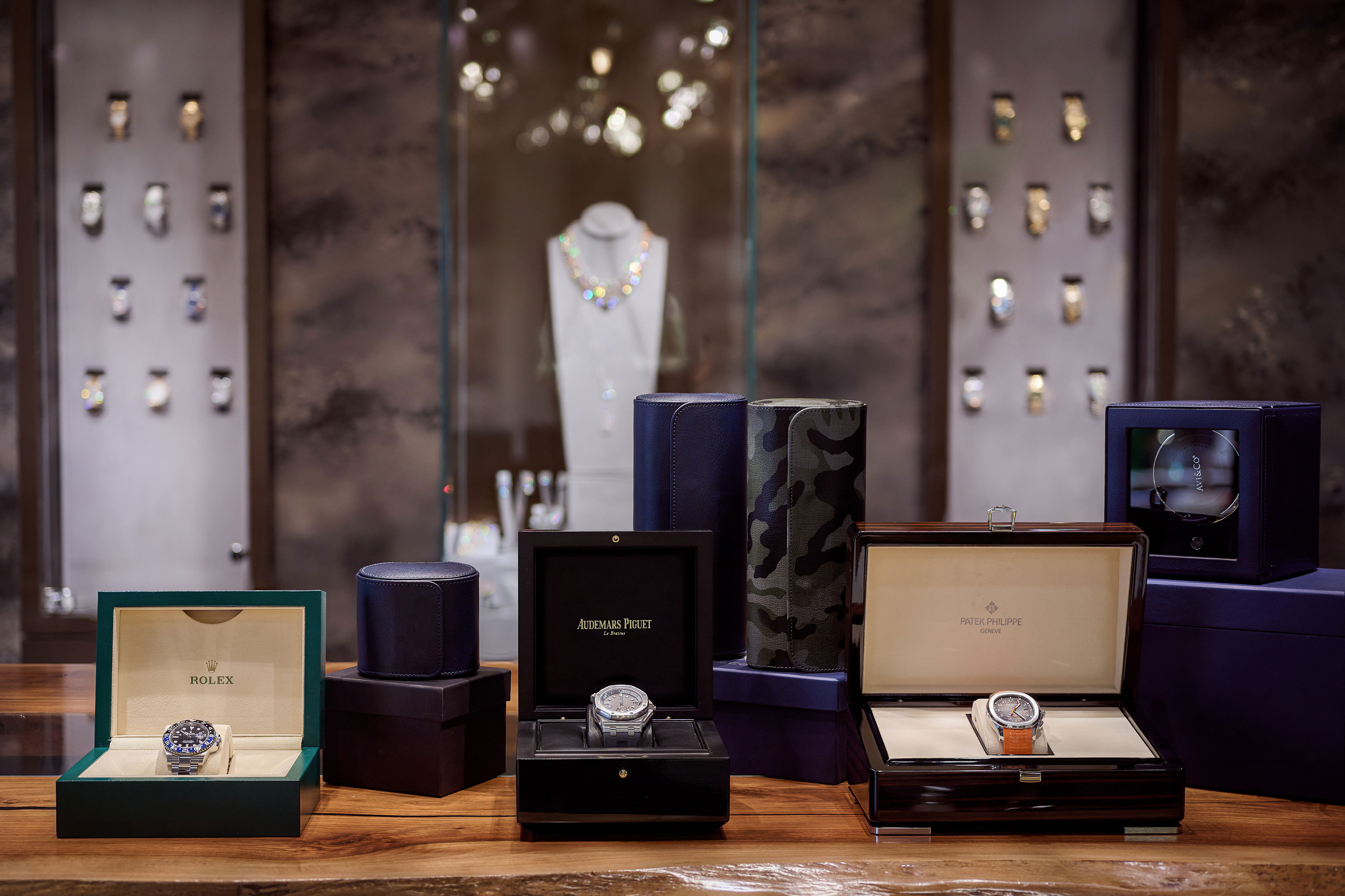 A Rolex, Audemars Piguet, and Patek Philippe Watch Sit in Their Original Brand Boxes Displayed in front of Watch Cases, Surrounded by a Watch Winder and Watch Rolls.