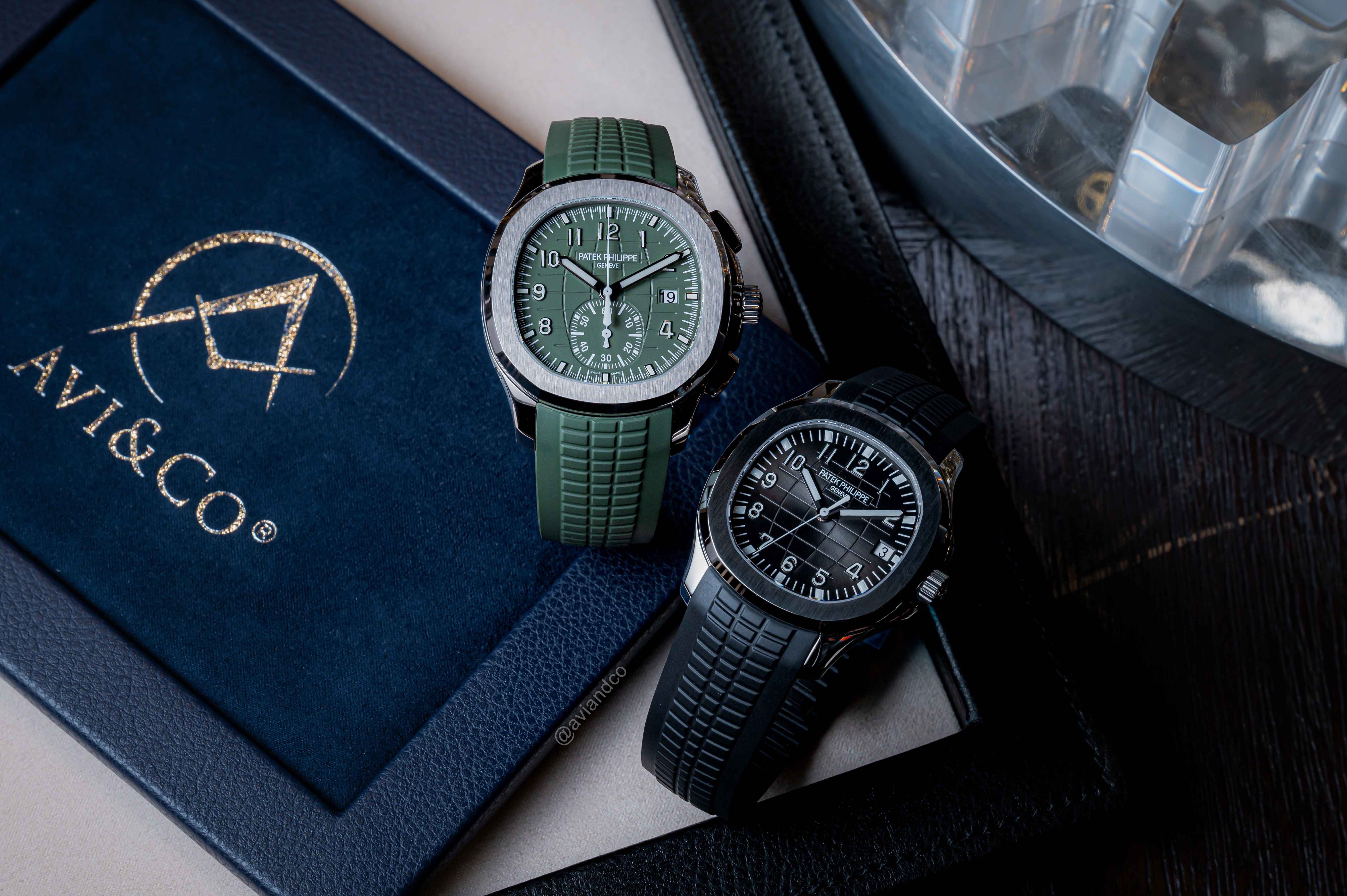 Two Similar Luxury Timepieces with Rubber Bracelets, Arabic Numerals, and Fixed Bezels with Different Materials Displayed on Clear C-Rings Above a Navy-Blue Avi & Co. Watch Pad.