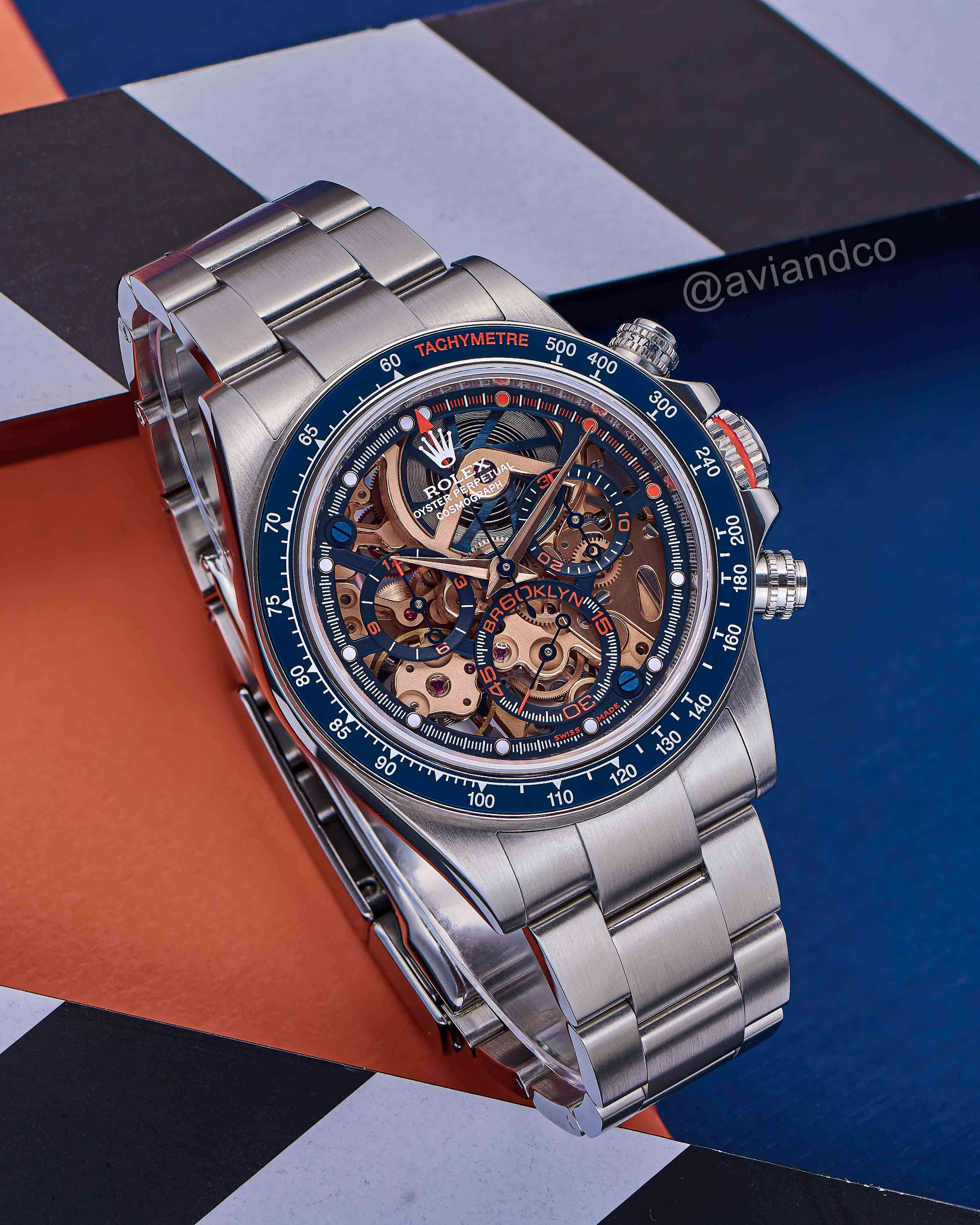 Openworked Dial with a Blue Bezel Timepiece, Sterling Silver Bracelet on a C-Ring on an Orange, Blue, Black, and White Background.