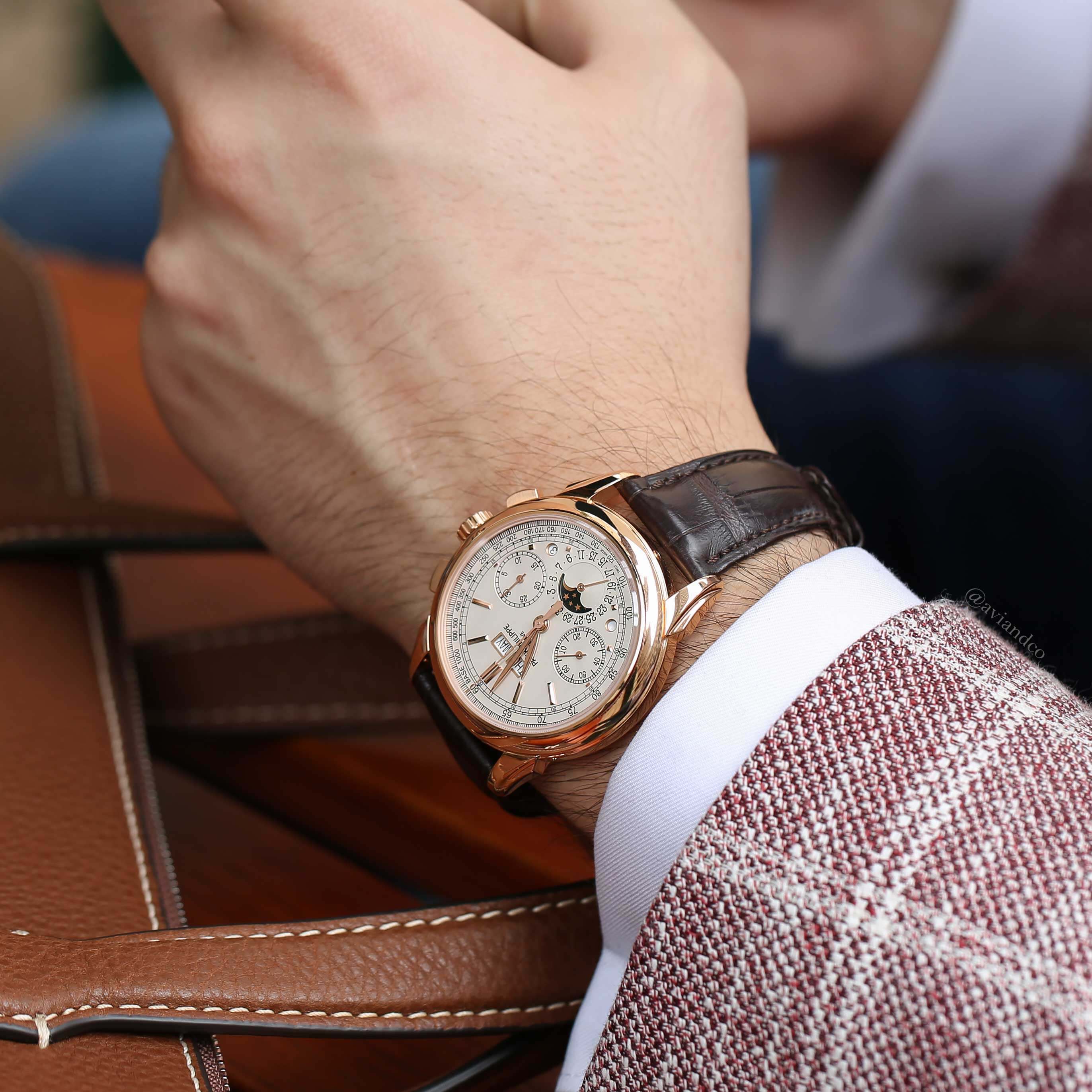 White Dial Timepiece with Brown Leather Strap on a Man’s Wrist Wearing a Pink Plaid Jacket Near a Brown Leather Briefcase.