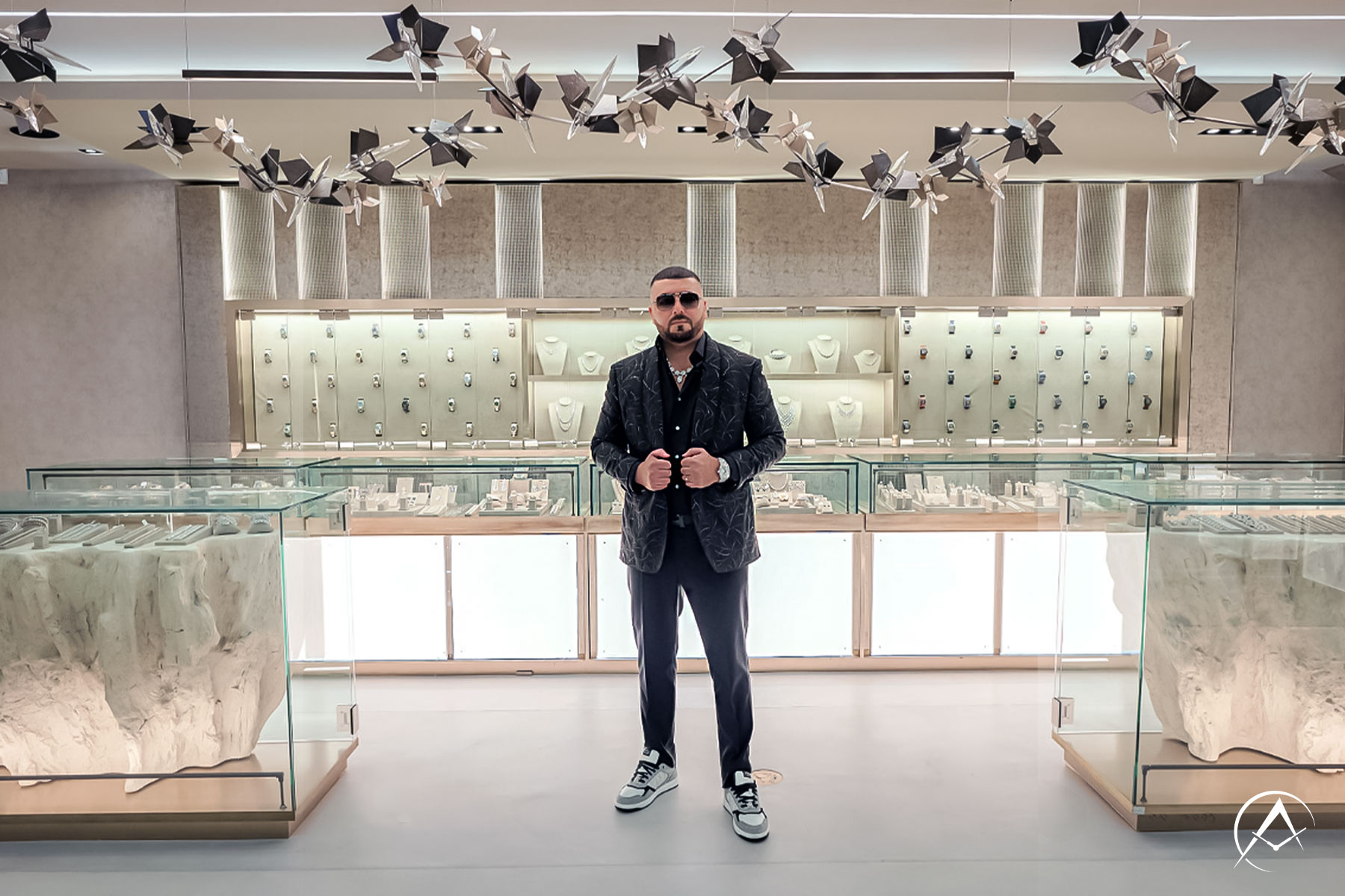 Avi Hiaeve Stands in the Middle of his Miami Showroom, Surrounded by Watches and Jewelry in Display Cases, Wearing a Black Suit, Avi & Co. Limited Edition Timepiece, and The Crown Chain.