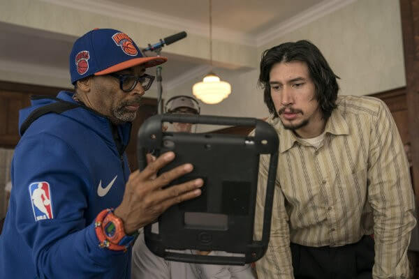 Spike Lee Wears a Blue Nike Sweatshirt, New York Knicks Baseball Cap, and Orange and Blue Timepiece. He Shares a Black Monitor with Actor Adam Driver, Who Wears a Beige Button-Down Shirt and Black Pants.