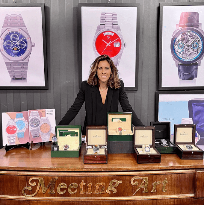 Giorgia Mondani Stands Behind a Wooden Desk which Displays Luxury Watches in their Boxes with Timepieces Framed on the Wall.