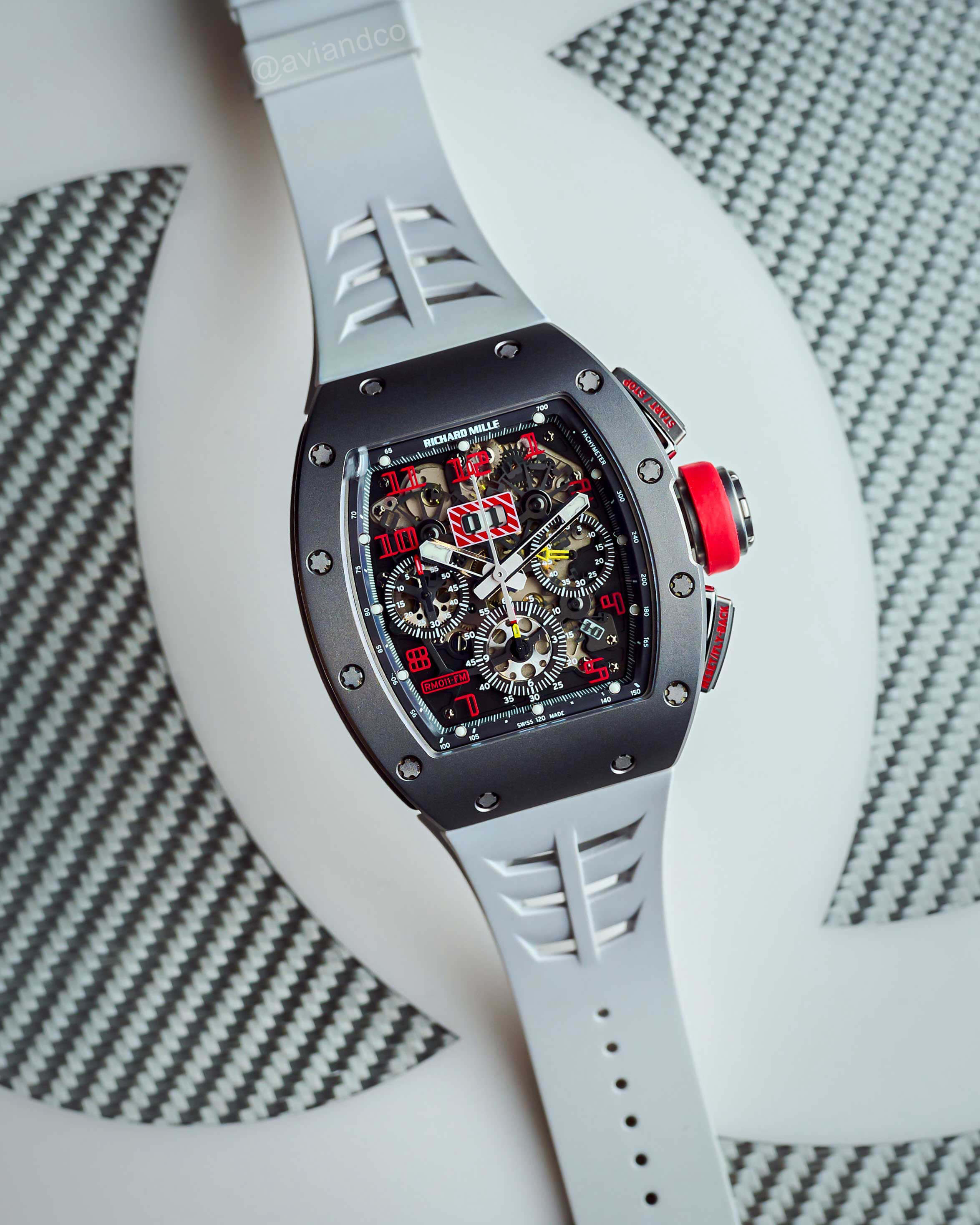 Openworked Dial Timepiece with Red Crown and Gray Rubber Strap Laid Out on a White and Gray Background.