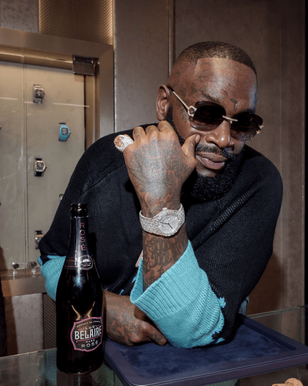 Man Wearing Sunglasses Poses with Diamond-Pave Timepiece and Diamond-Pave Pinky Ring in Front of Richard Mille Timepieces in Glass Case.