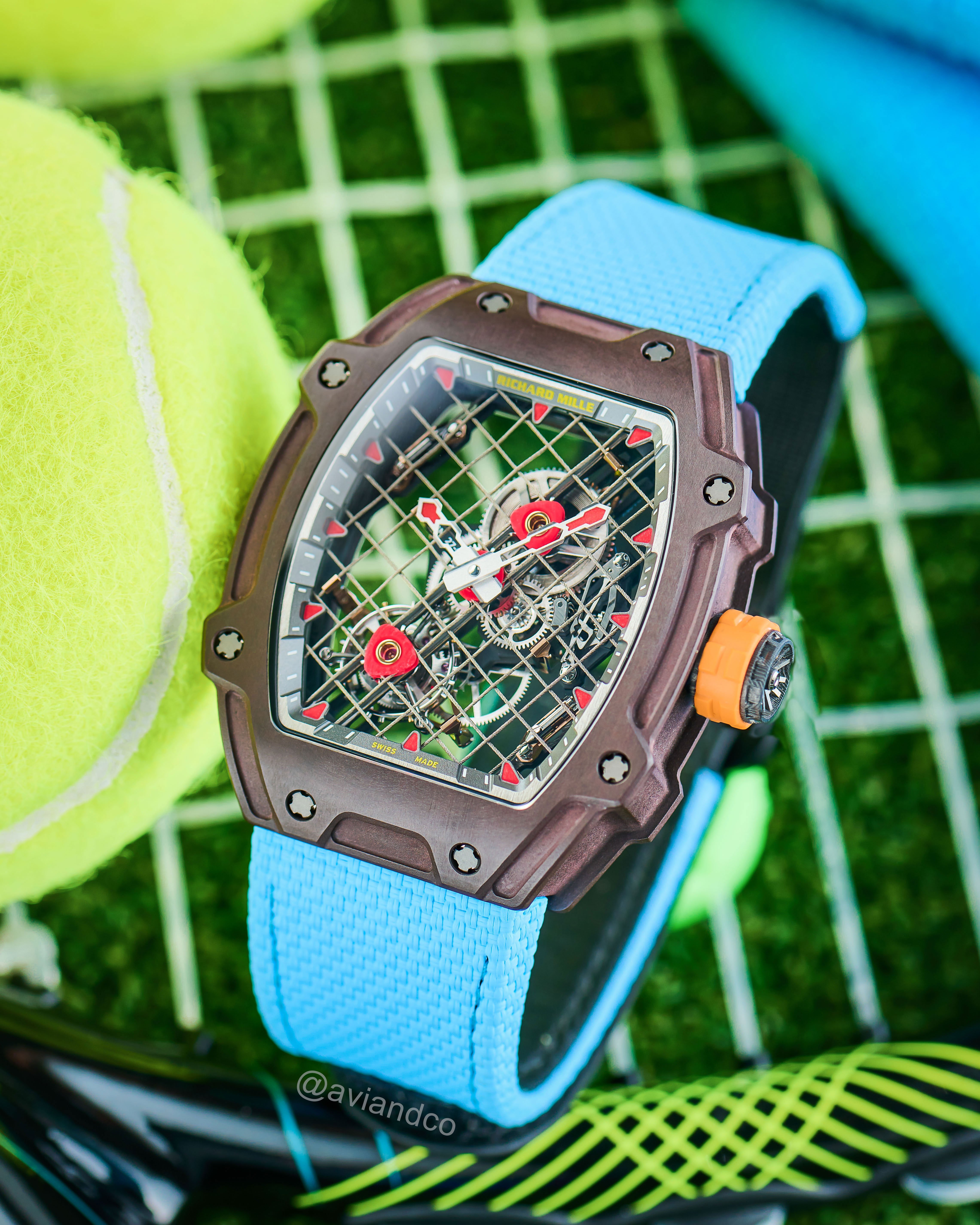 Openworked Dial Timepiece with Blue Strap and Orange Crown Displayed on a Tennis Racket Beside a Tennis Ball.
