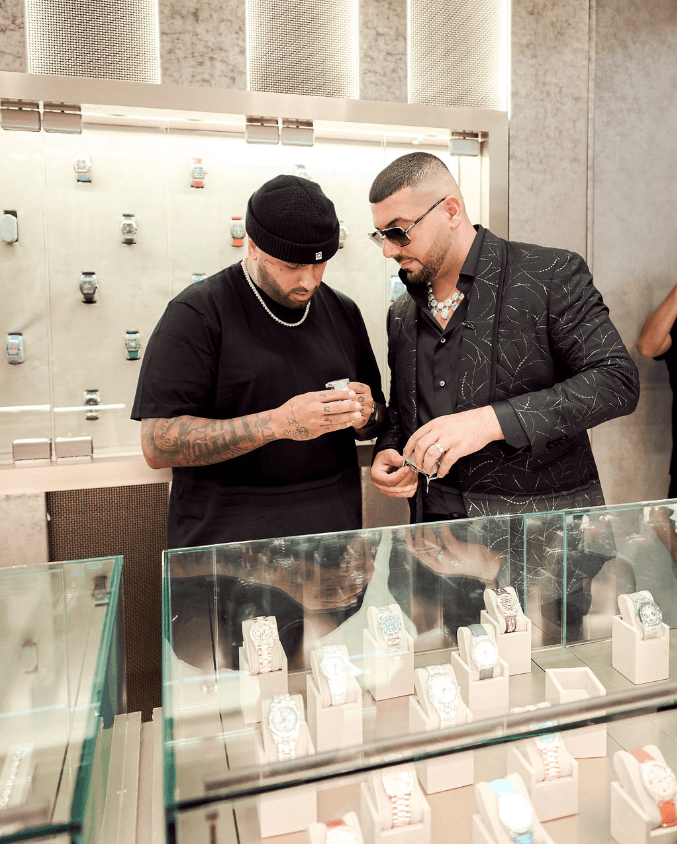 Man Wearing Black T-shirt and Hat Holds Avi & Co. Limited Edition Diamond-Pave Timepiece Beside Avi Hiaeve Between Glass Display Cases Filled with Luxury Watch Brands Such as Richard Mille, Patek Philippe, Audemars Piguet, and Rolex.
