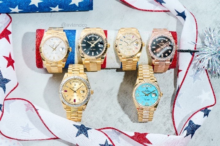 Six Luxury Rolex Timepieces Sit on C-Rings on a White Marble Tabletop with Red, White, and Blue Ribbon Surrounding Them.