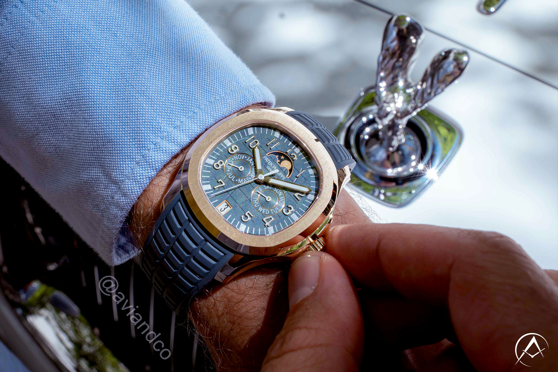Man Wrist Wearing 18K Rose Gold Luxury Timepiece with Blue Dial, Arabic Numerals, Navy Blue Rubber Strap, Annual Calendar, and Date Function in Front of Silver Luxury Car.